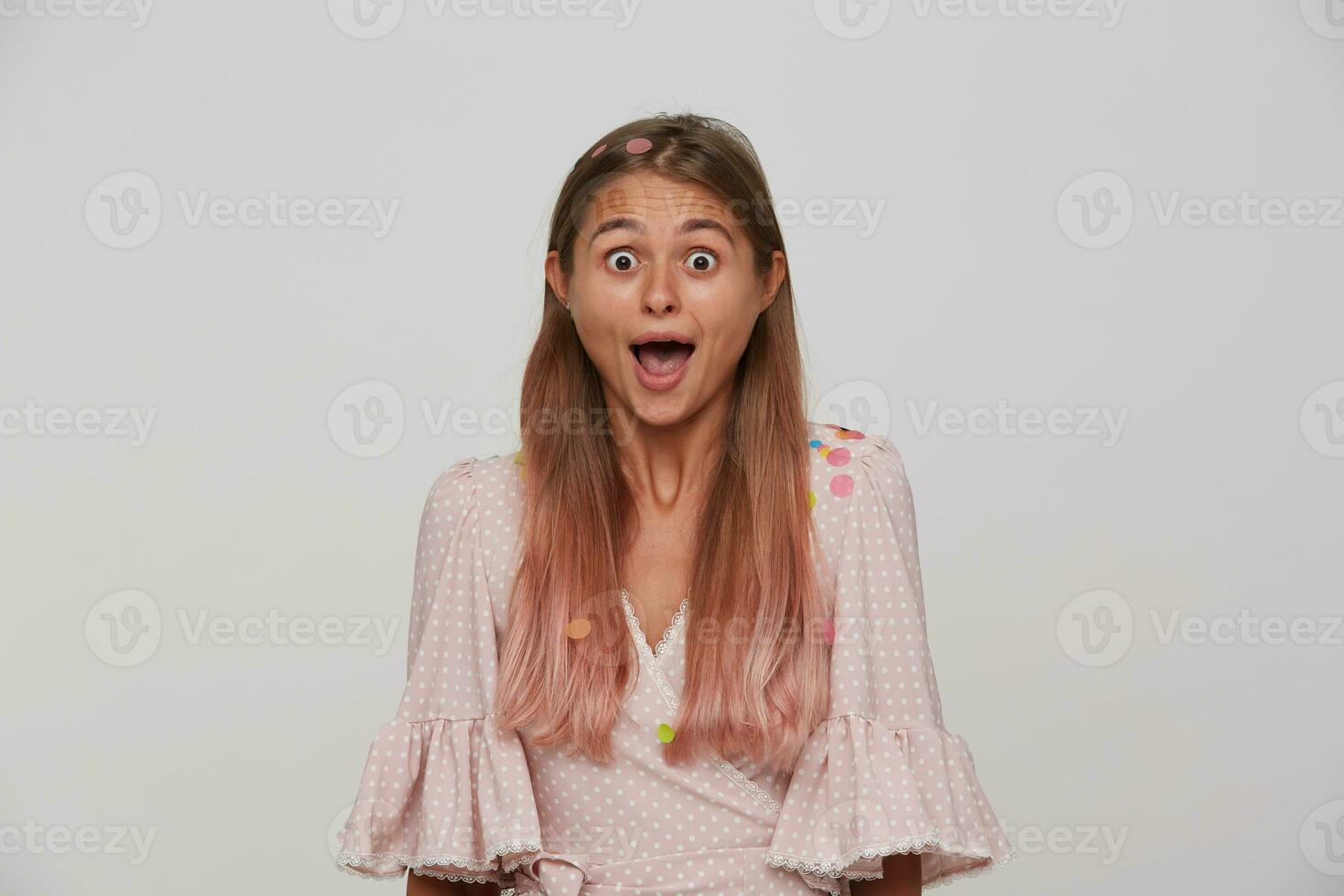 interior foto de hermosa de ojos marrones joven largo peludo rubia dama mirando sorprendentemente a cámara mientras en pie terminado blanco antecedentes en rosado romántico vestir