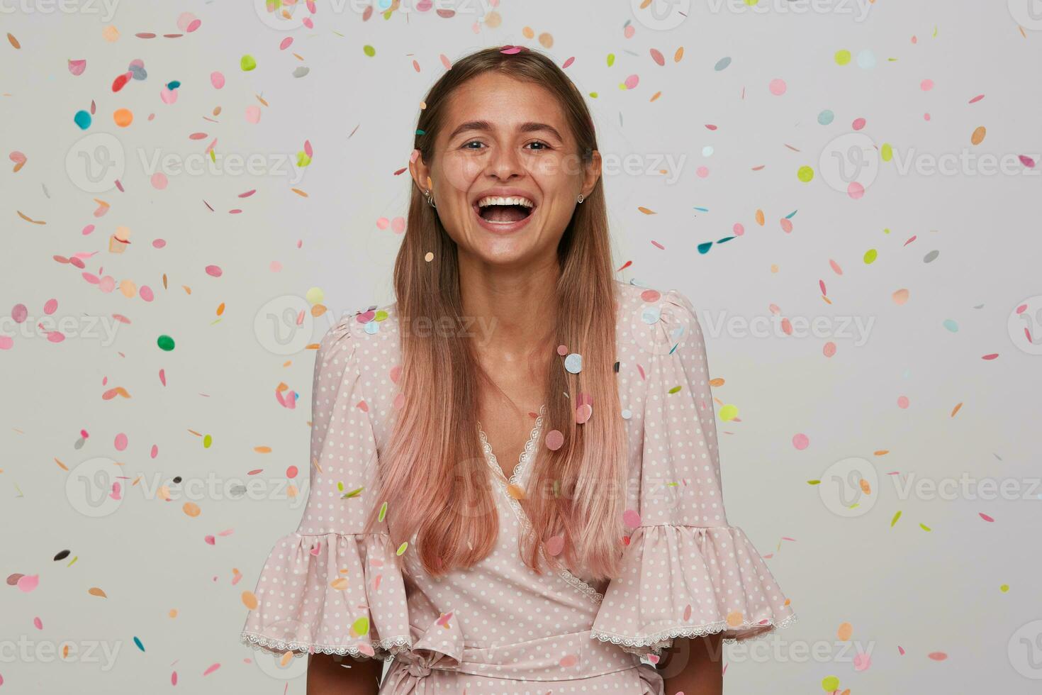 Studio shot of lovely happy young long haired lady laughing cheerfully while having birthday party with her friends, standing over white background in multicolored confetti photo