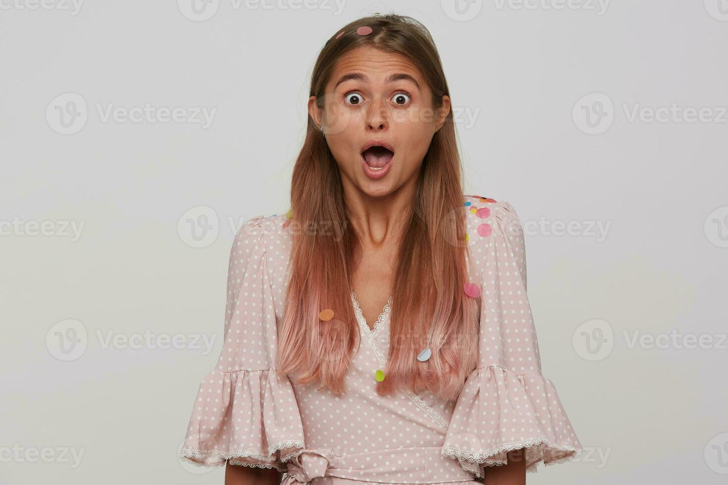 Astonished young lovely blonde female with long hair looking at camera with wide eyes and mouth opened, being amazed about something while posing over white background photo
