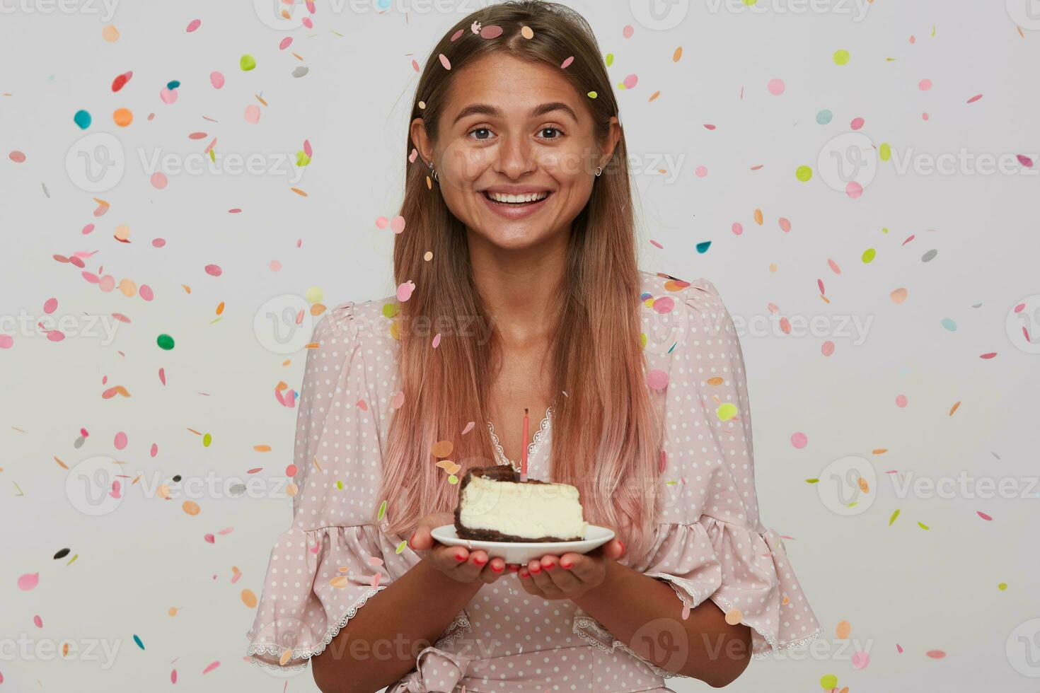 Studio shot of cheerful young lovely lady with light brown long hair holding plate with birthday cake and looking happily to camera with wide smile, isolated over white background photo