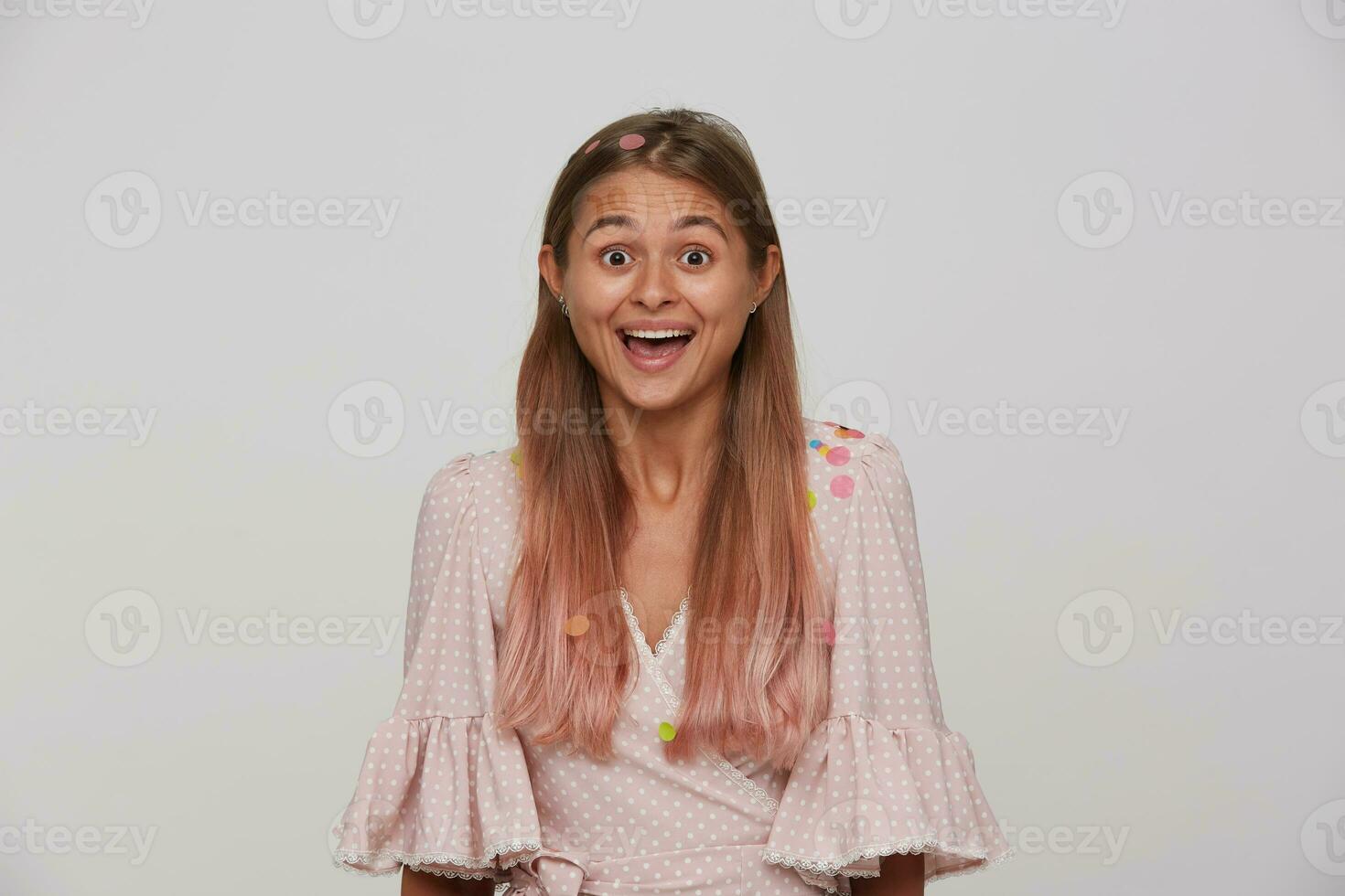 Surprised young pretty brown-eyed long haired woman in festive elegant clothes posing over white background, looking joyfully at camera with wide mouth opened photo
