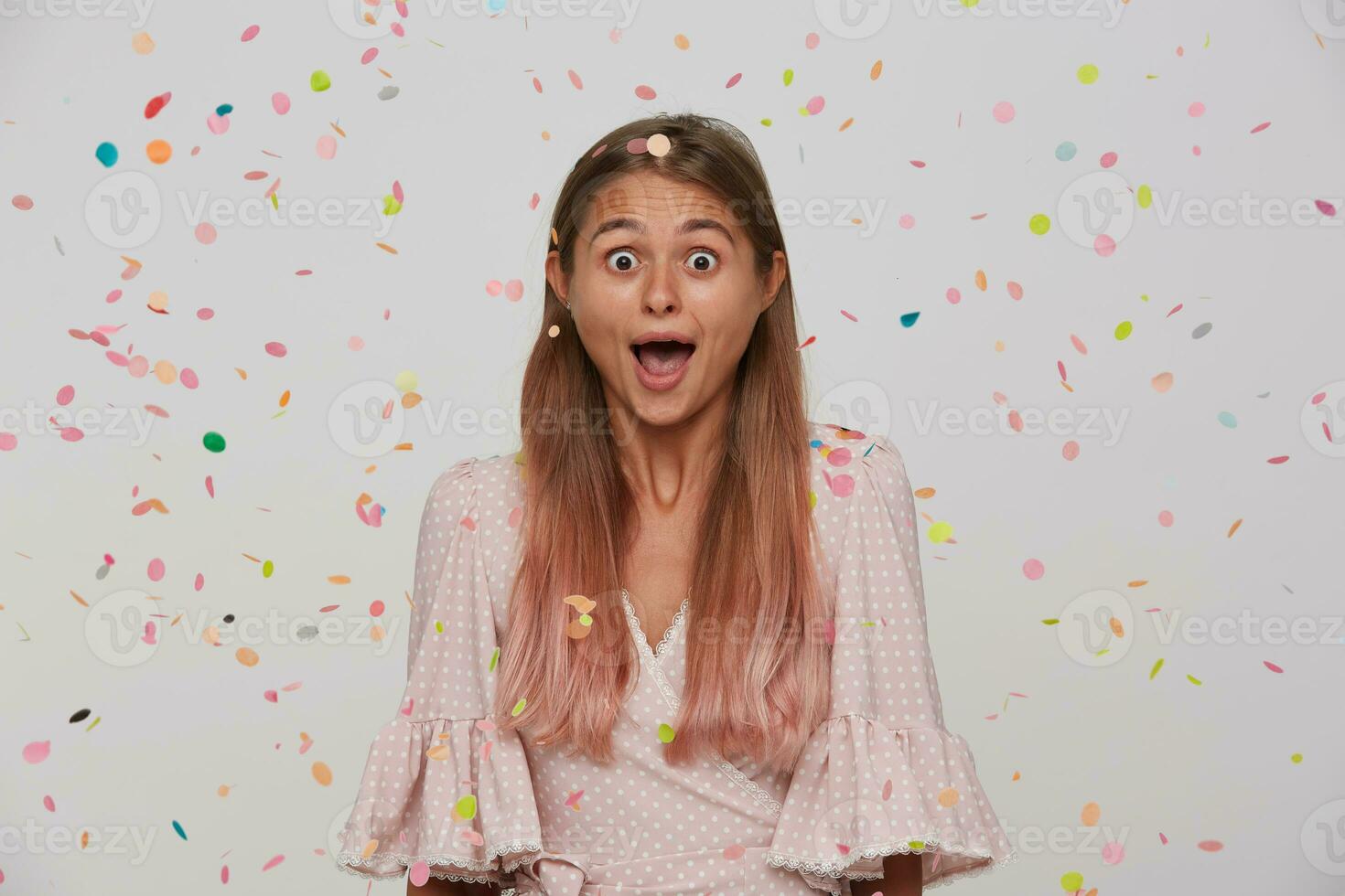 Open-eyed young attractive woman with light brown long hair wearing natural makeup while standing over white background and multicolored confetti, looking surprisedly at camera with opened mouth photo