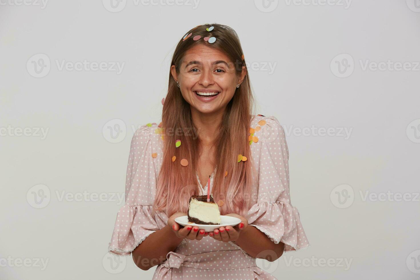 Positive young lady with light brown long hair smiling pleasantly at camera while rejoicing about birthday party with her friends, keeping piece of cake in hands while posing over white background photo