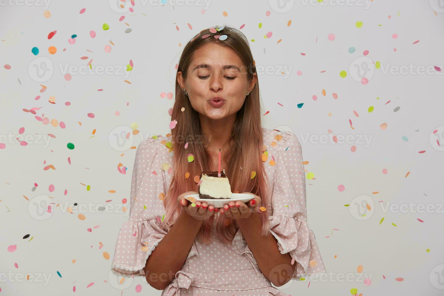 Indoor shot of lovely young long haired blonde woman blowing out candle on birthday cake while standing over white background, dressed in pink romantic dress photo