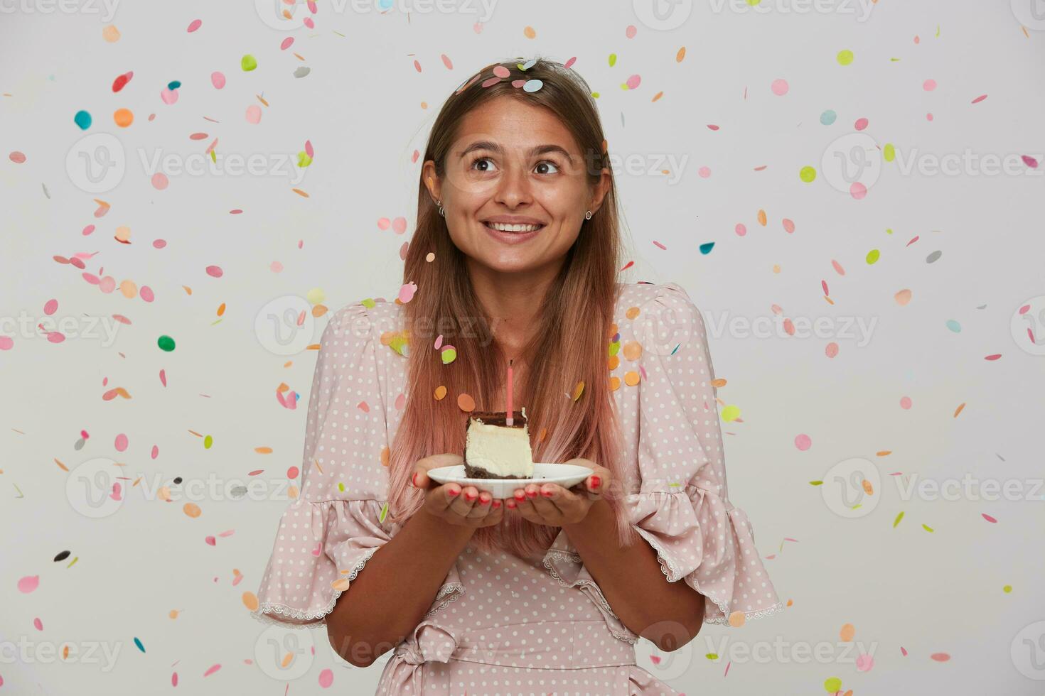 Surprised young brown-eyed lovely long haired blonde woman with natural makeup having birthday party and keeping piece of cake in raised hands while posing over white background photo