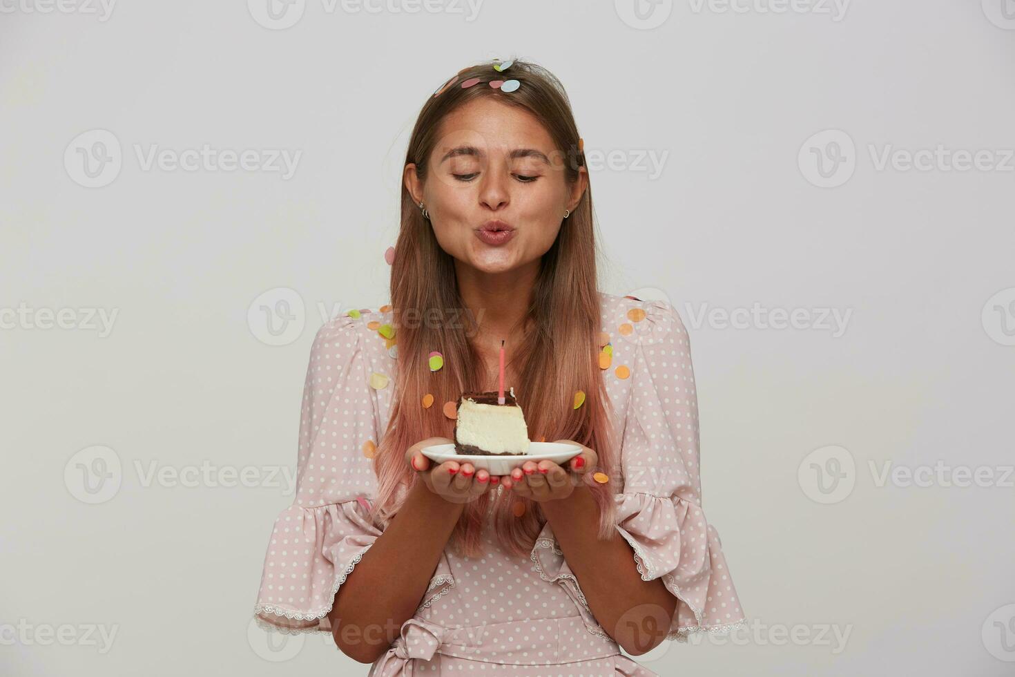 Positive young attractive long haired blonde lady blowing on candle while holding birthday cake, celebrating birthday with her family and being in nice mood while posing over white background photo