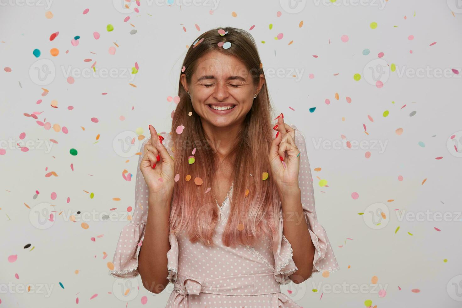 Pleasant looking young happy long haired blonde lady smiling happily while making wish on her birthday and keeping eyes closed, wearing pink romantic dress while standing over white background photo