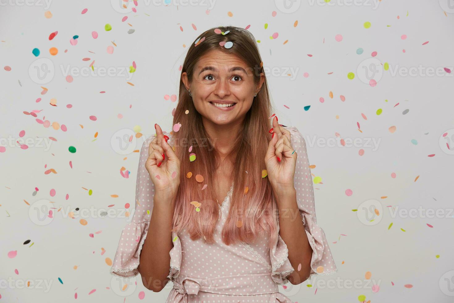 Positive young lovely brown-eyed long haired woman biting worringly underlip while making wish with crossed finfers, posing over white background in multicolored confetti photo