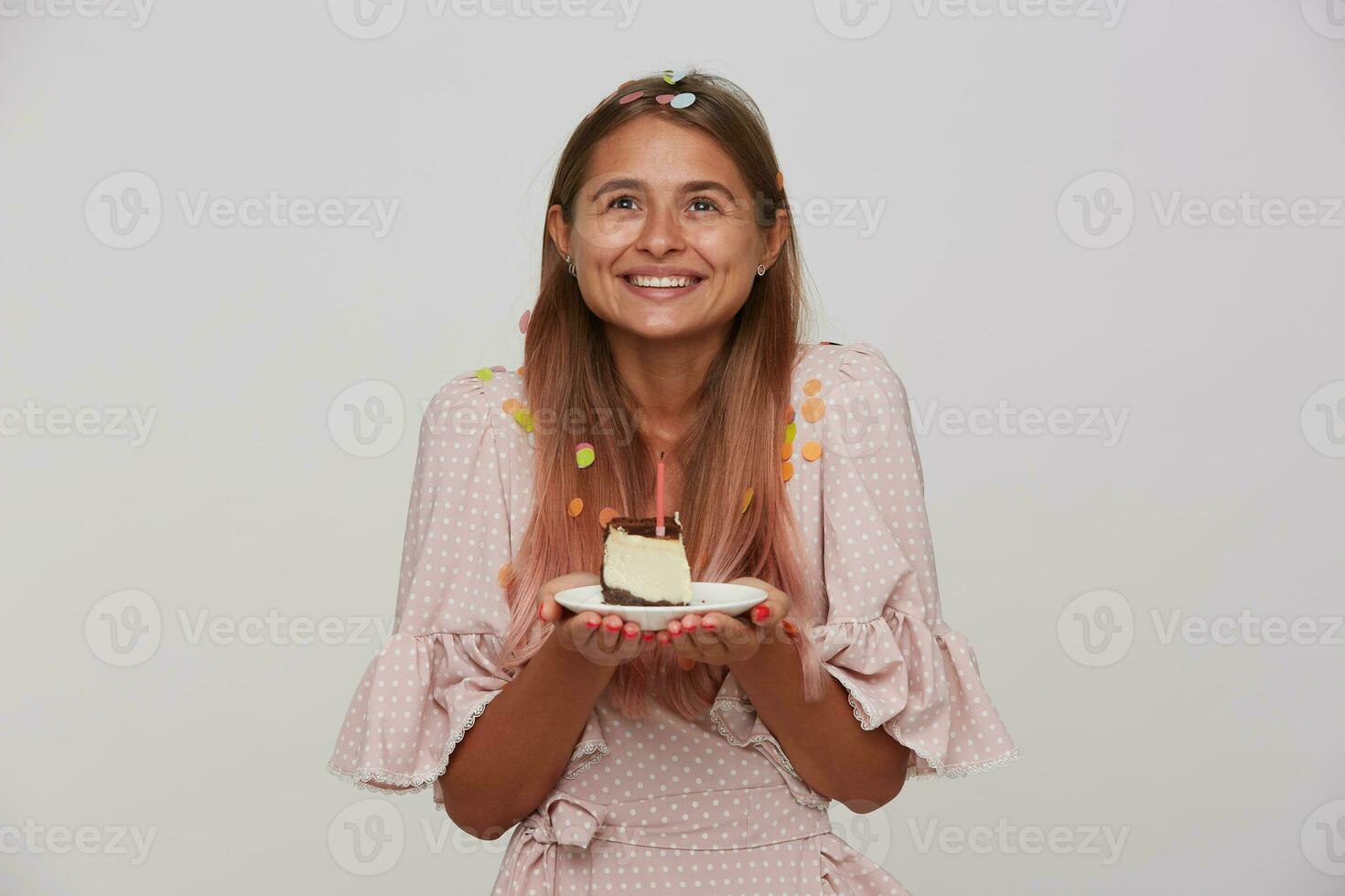 Indoor shot of positive young long haired blonde female dressed in pink romantic dress smiling happily and keeping plate with birthday cake, standing over white background photo