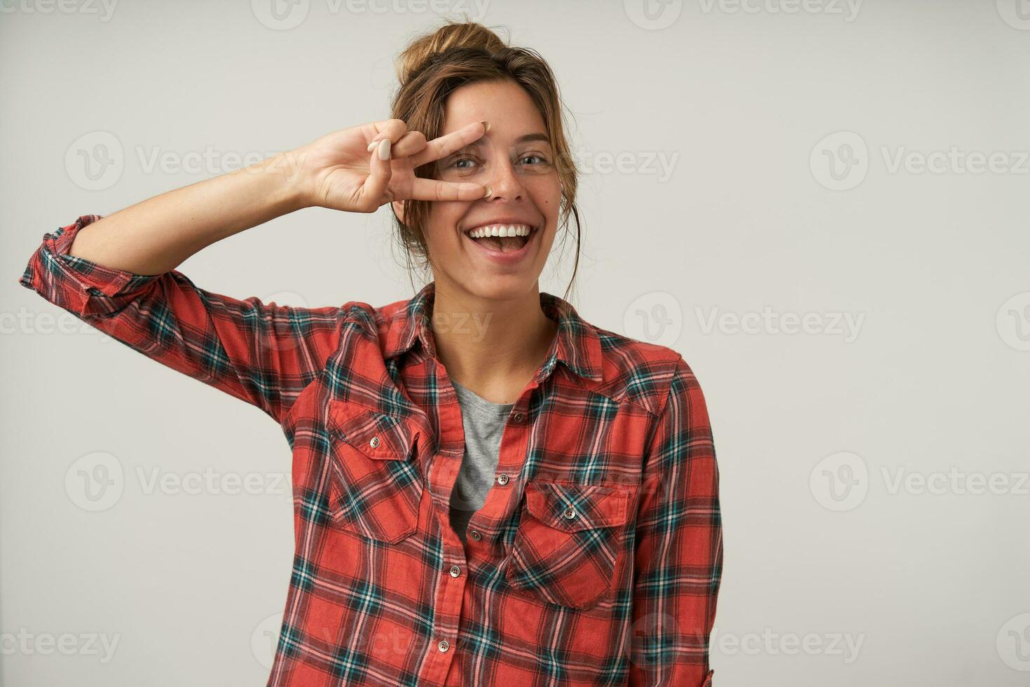 Portrait of young happy brunette woman keeping peace gesture near her face while looking joyfully at camera, standing over white background in casual wear photo