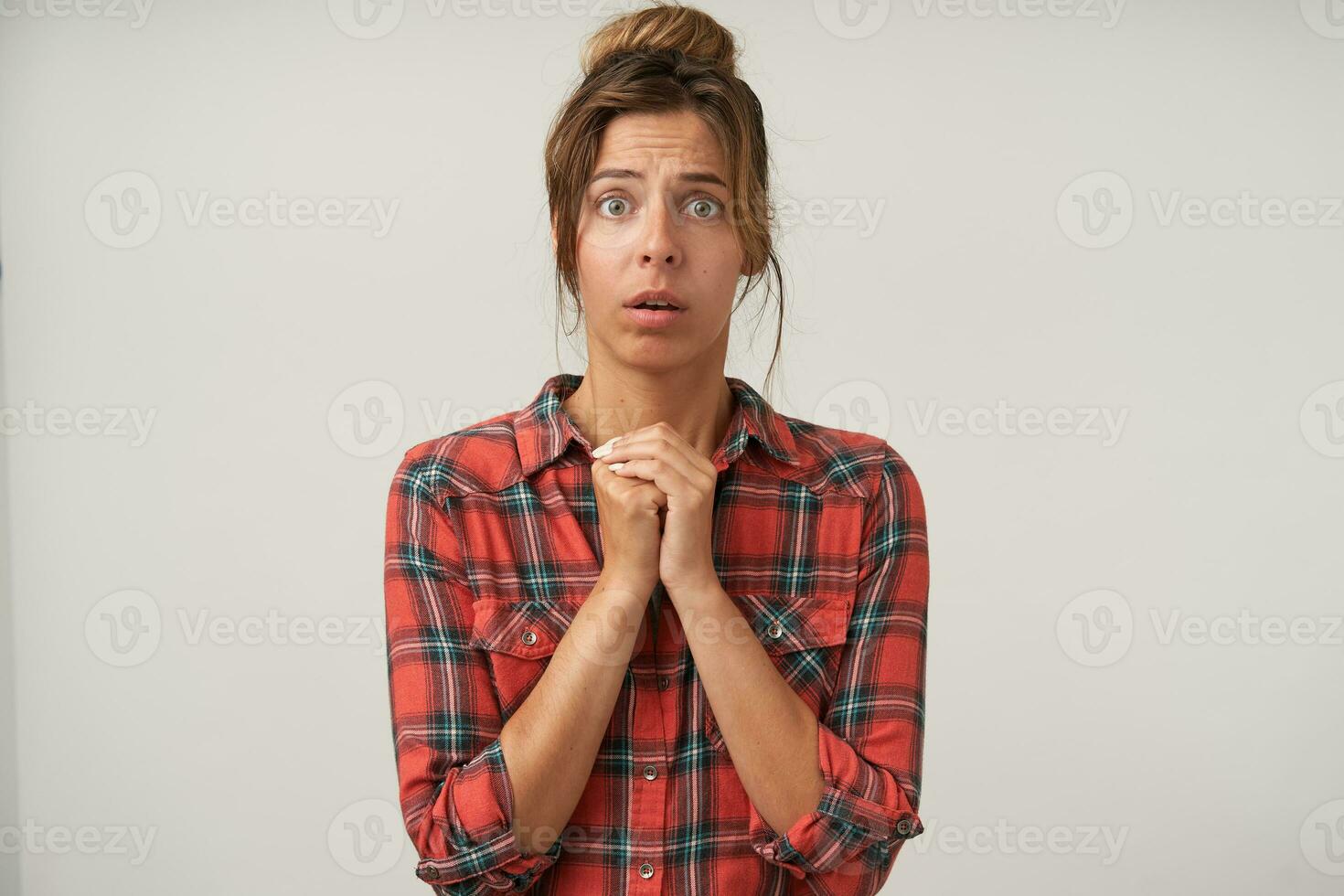 Surprised young pretty brunette female dressed in checkered shirt keeping folded hands under her chin while looking worringly at camera, isolated over white background photo