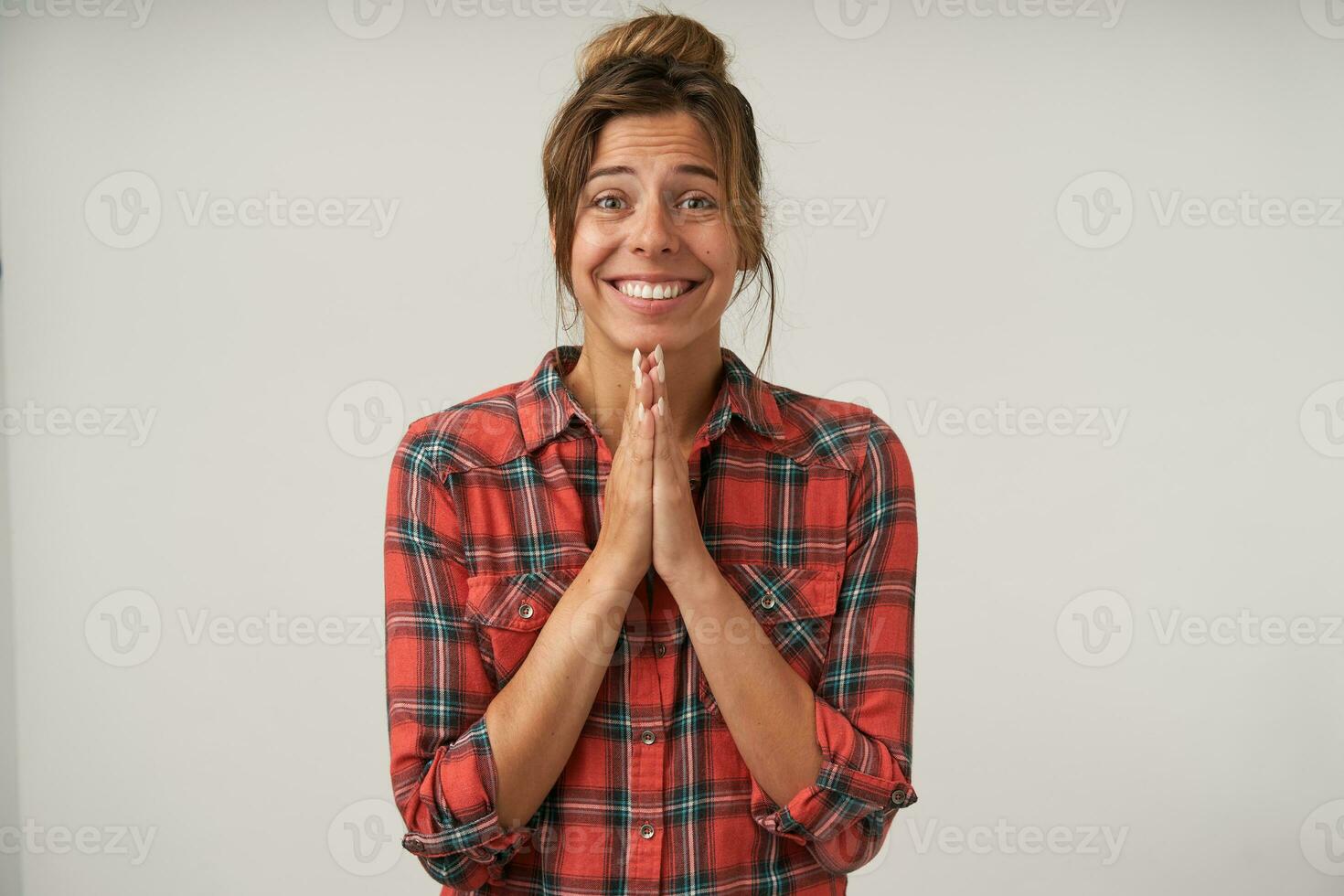 Indoor shot of young attractive brown haired lady with natural makeup keeping raised palms together while looking hopefully at camera, posing over white background photo