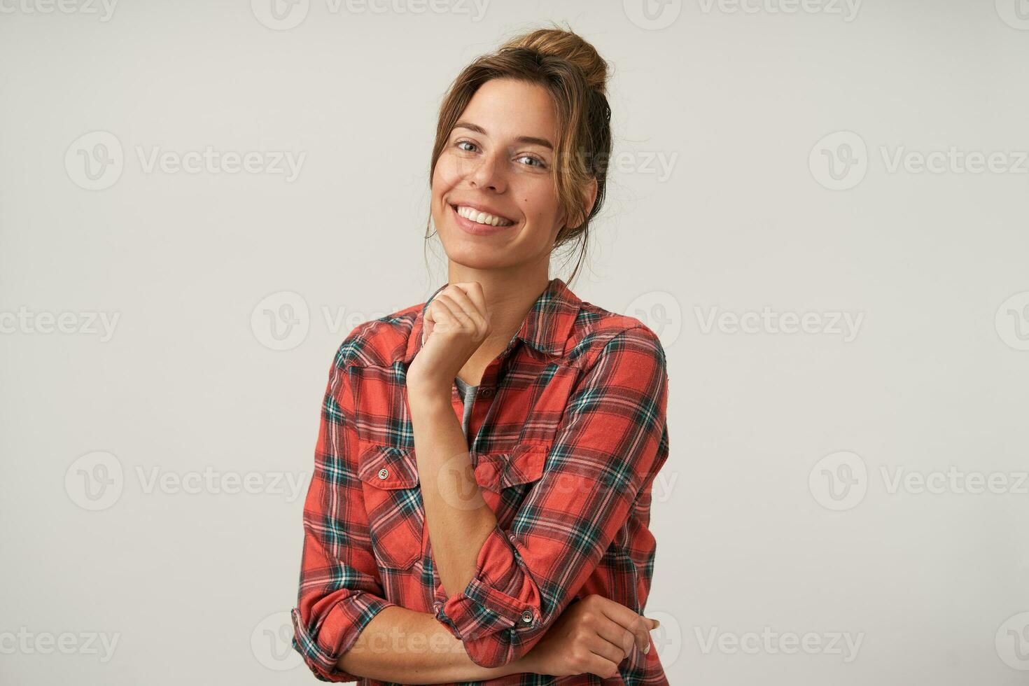 Charming young lovely brown haired woman with natural makeup leaning her chin on raised hand while looking cheerfully at camera with wide smile, posing over white background photo
