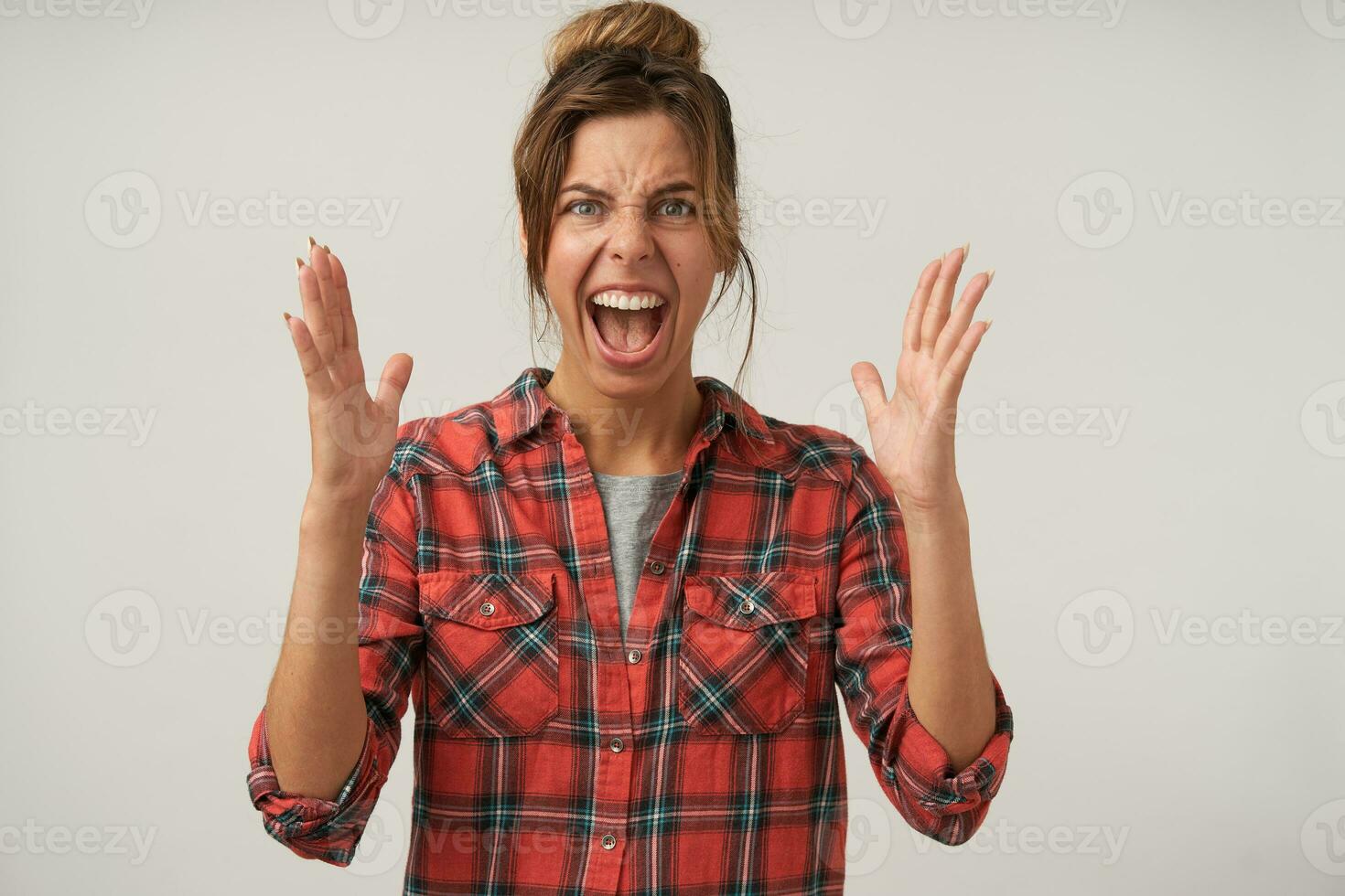 Portrait of expressive young brown haired woman with casual hairstyle frowning her face while screaming and raising emotionally hands, standing over white background photo