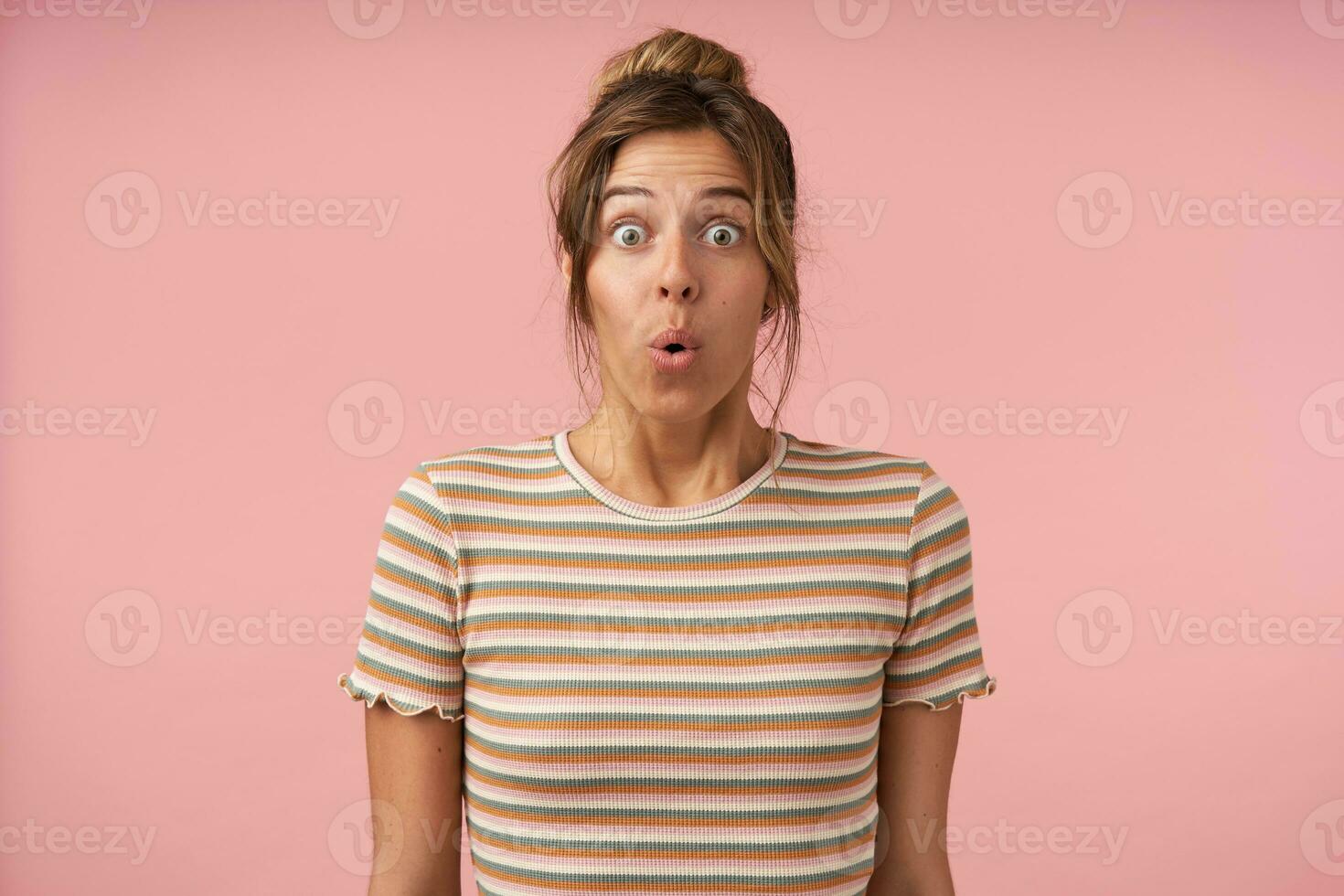Dazed young pretty brown haired woman with bun hairstyle rounding amazedly her eyes while looking surprisedly at camera, isolated over pink background photo