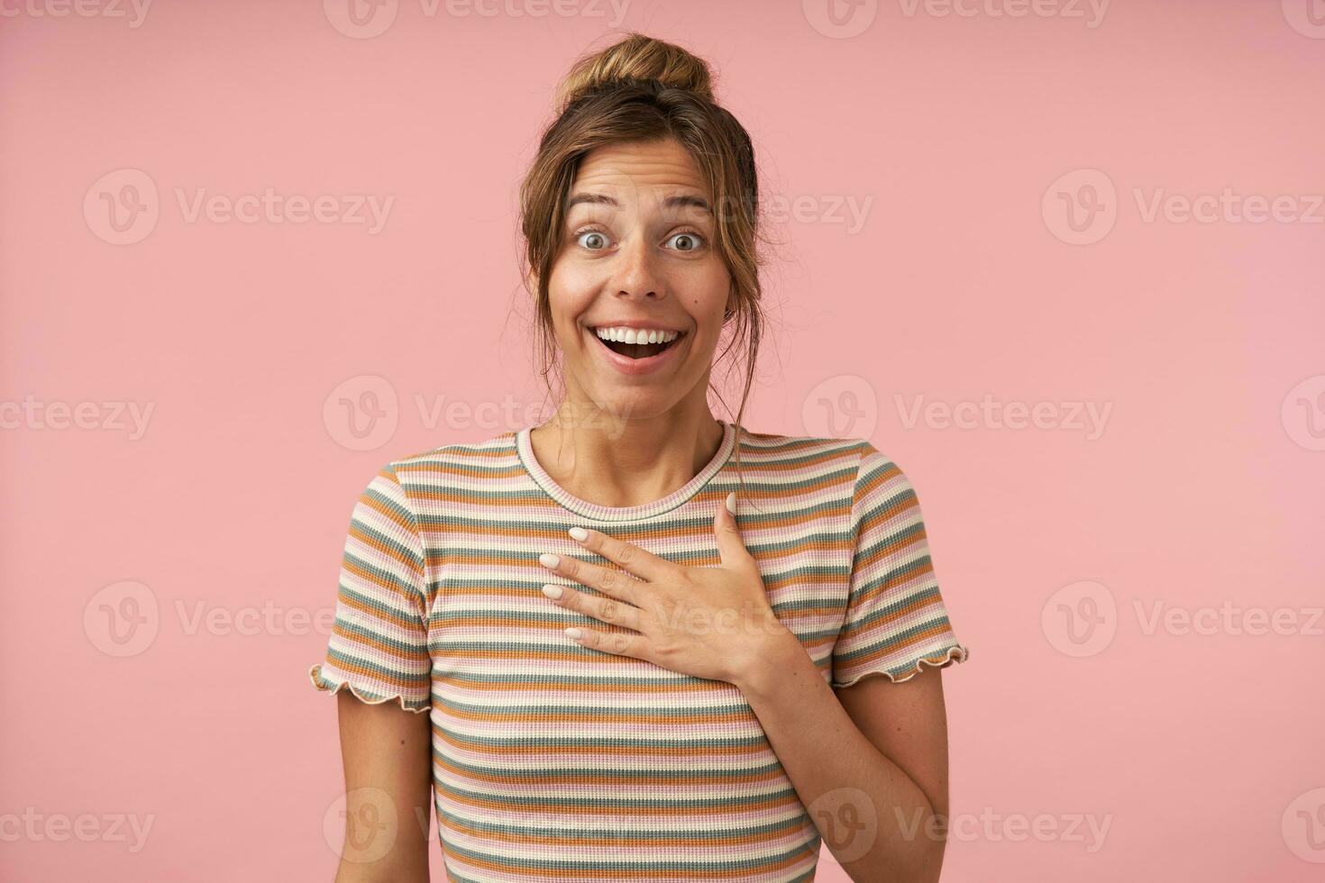 Excited young attractive brown haired lady raising surprisedly eyebrows while looking joyfully at camera and keeping palm on her chest, isolated over pink background photo