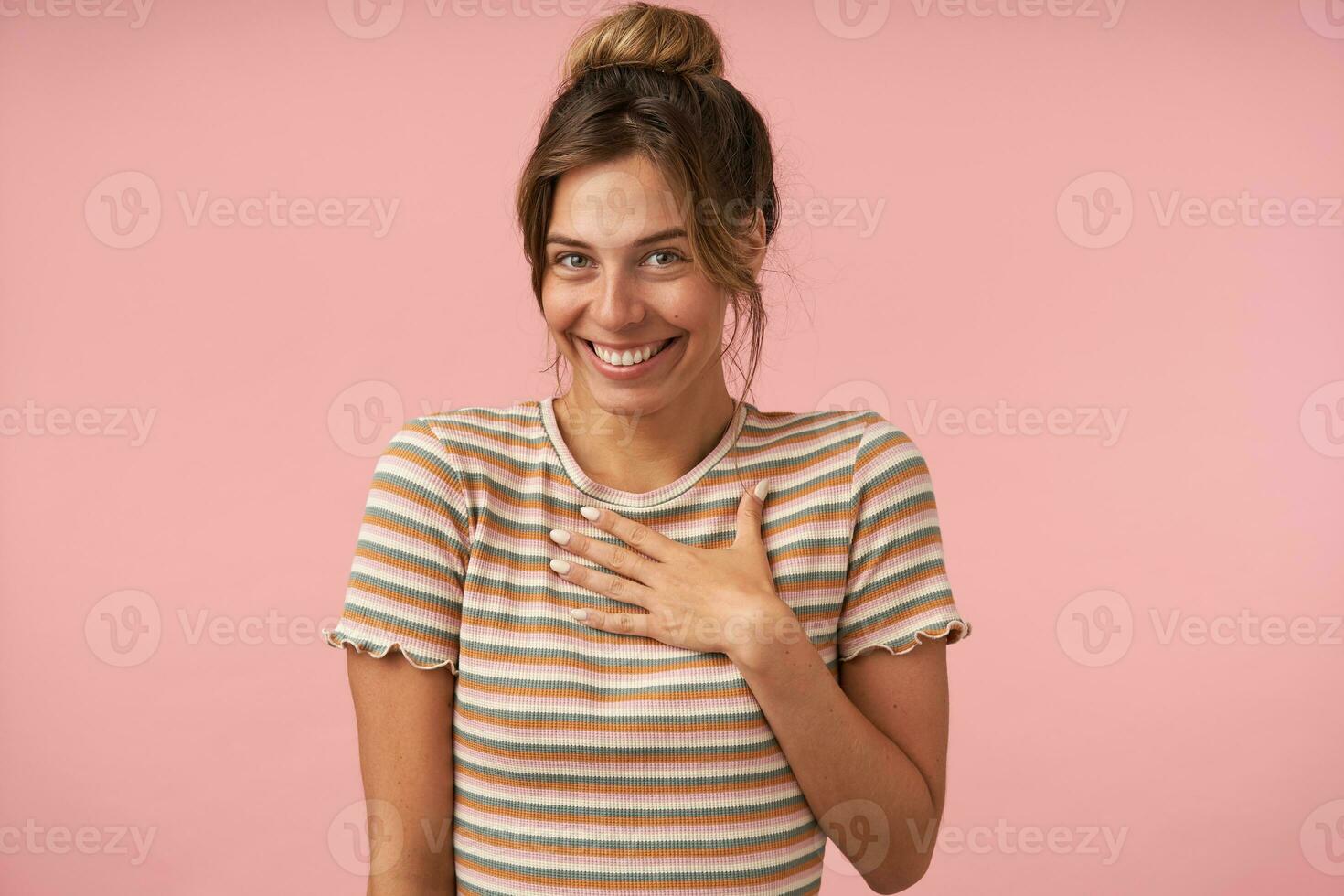 Portrait of young beautiful brown haired woman with natural makeup looking cheerfully at camera with wide smile while posing over pink background in casual wear photo
