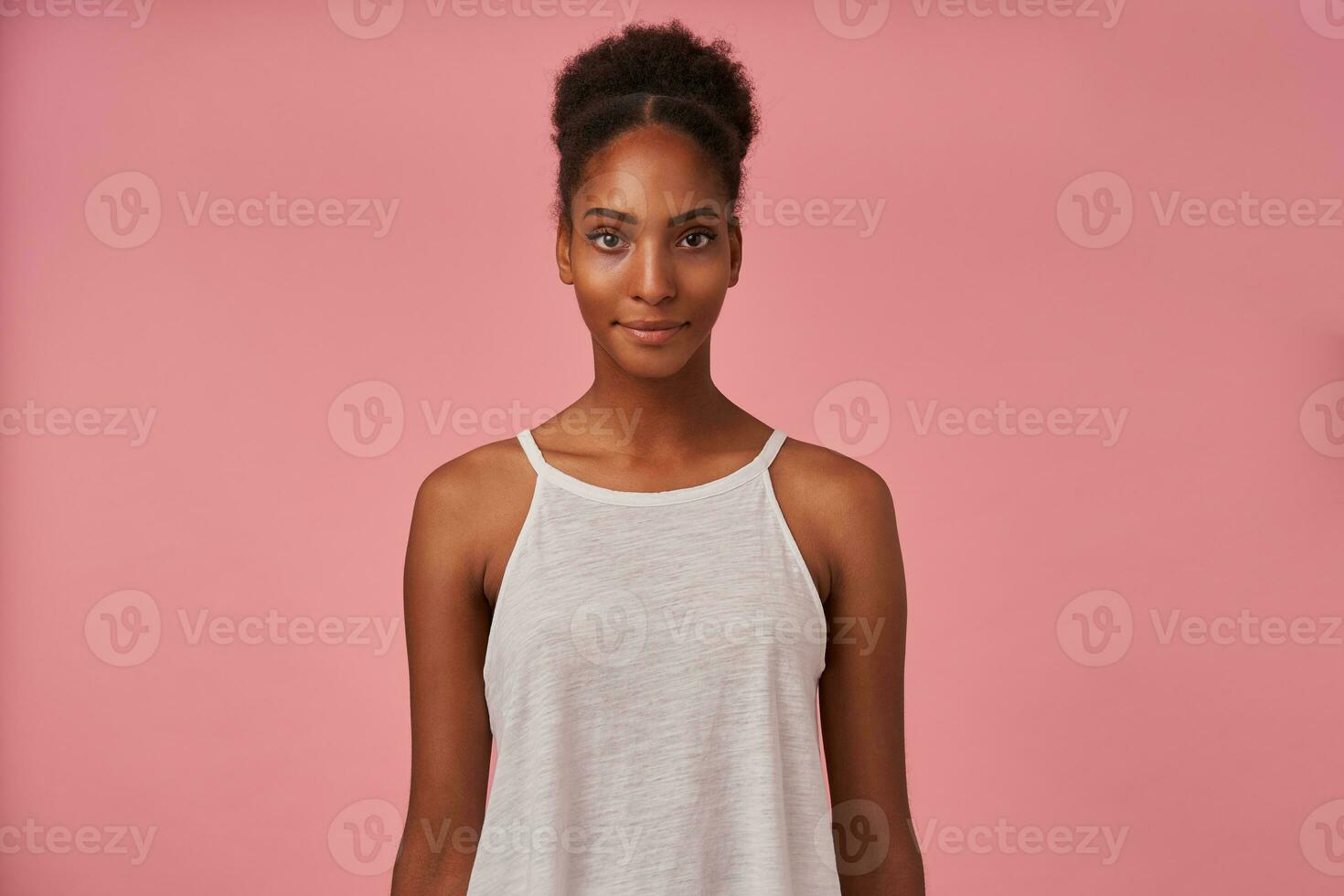 Portrait of young pretty brown-eyed brunette dark skinned woman keeping her lips folded while looking at camera with calm face, isolated over pink background photo