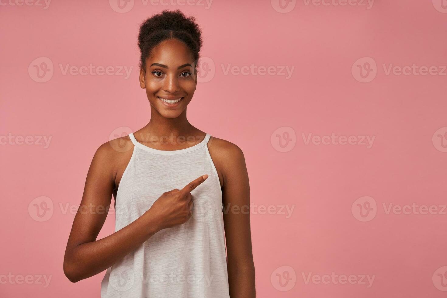 Pleased young lovely brown haired dark skinned female with bun hairstyle smiling ceerfully while pointing aside with forefinger, standing over pink background photo