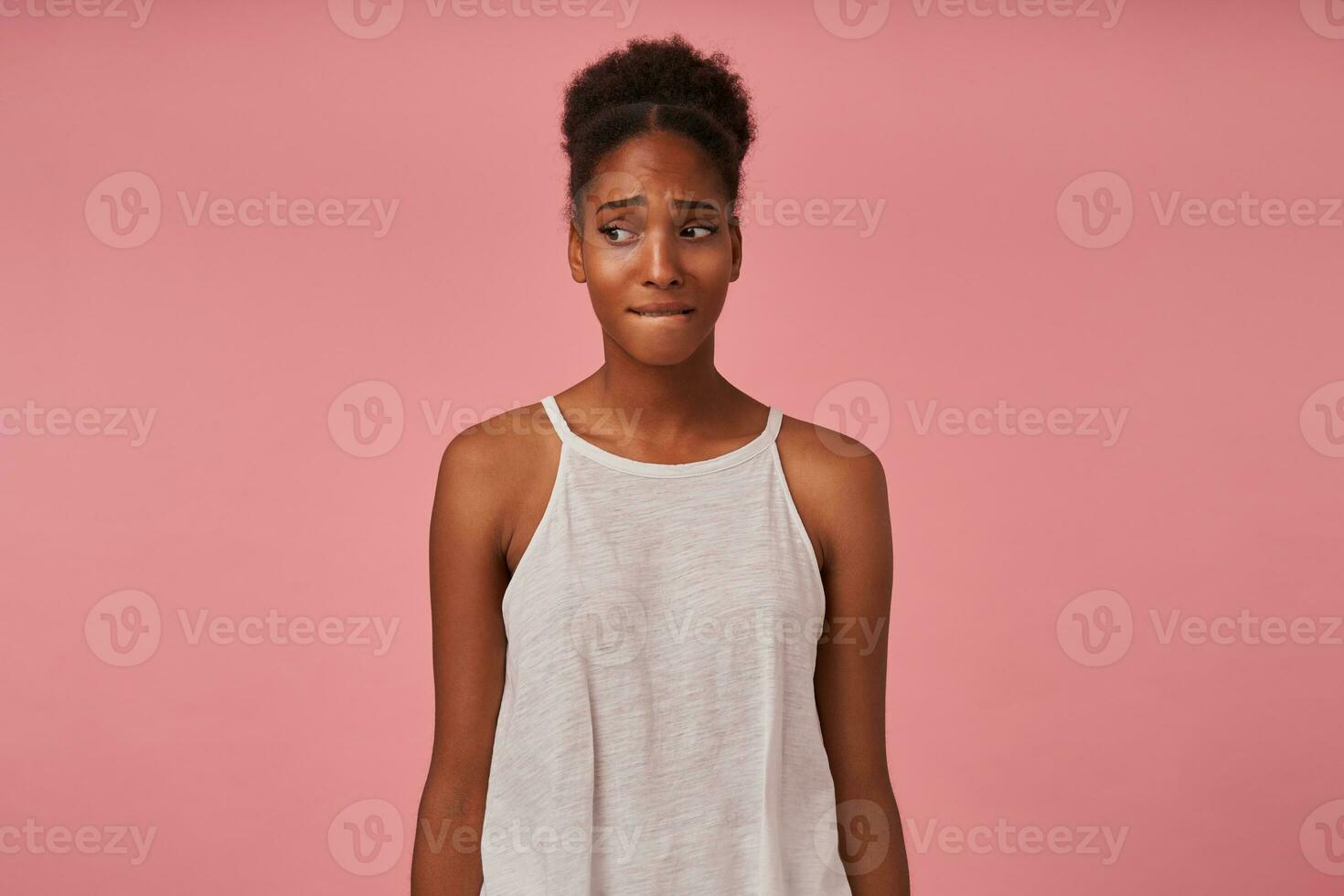 Frightened young curly brunette dark skinned lady biting her underlip and frowning eyebrows while looking sadly aside, isolated over pink background with hands down photo