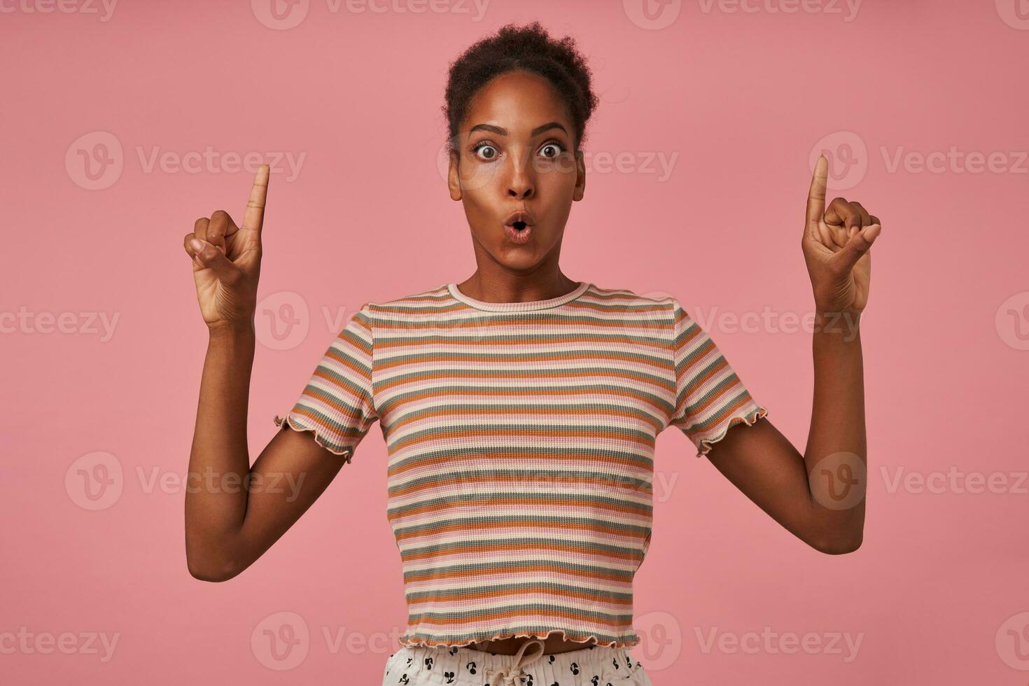 Bemused young pretty brown haired curly dark skinned woman rounding amazedly her eyes while showing upwards with raised index fingers, isolated over pink background photo