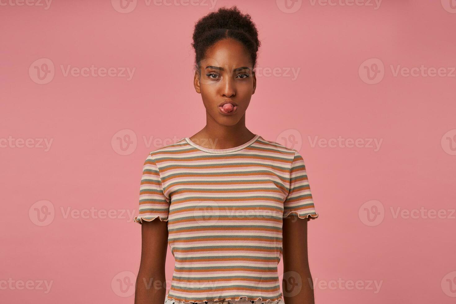 Indoor photo of young brown haired curly female with bun hairstyle showing tongue while looking at camera, dressed in beige t-shirt while posing over pink background