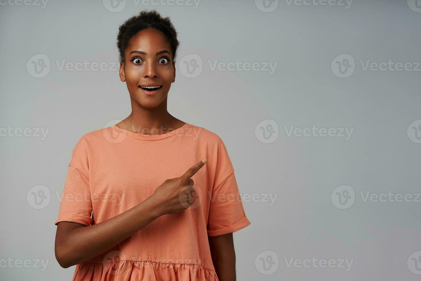 Agitated young lovely curly brunette woman with dark skin looking at camera with wide eyes opened while showing aside with index finger, isolated over grey background photo