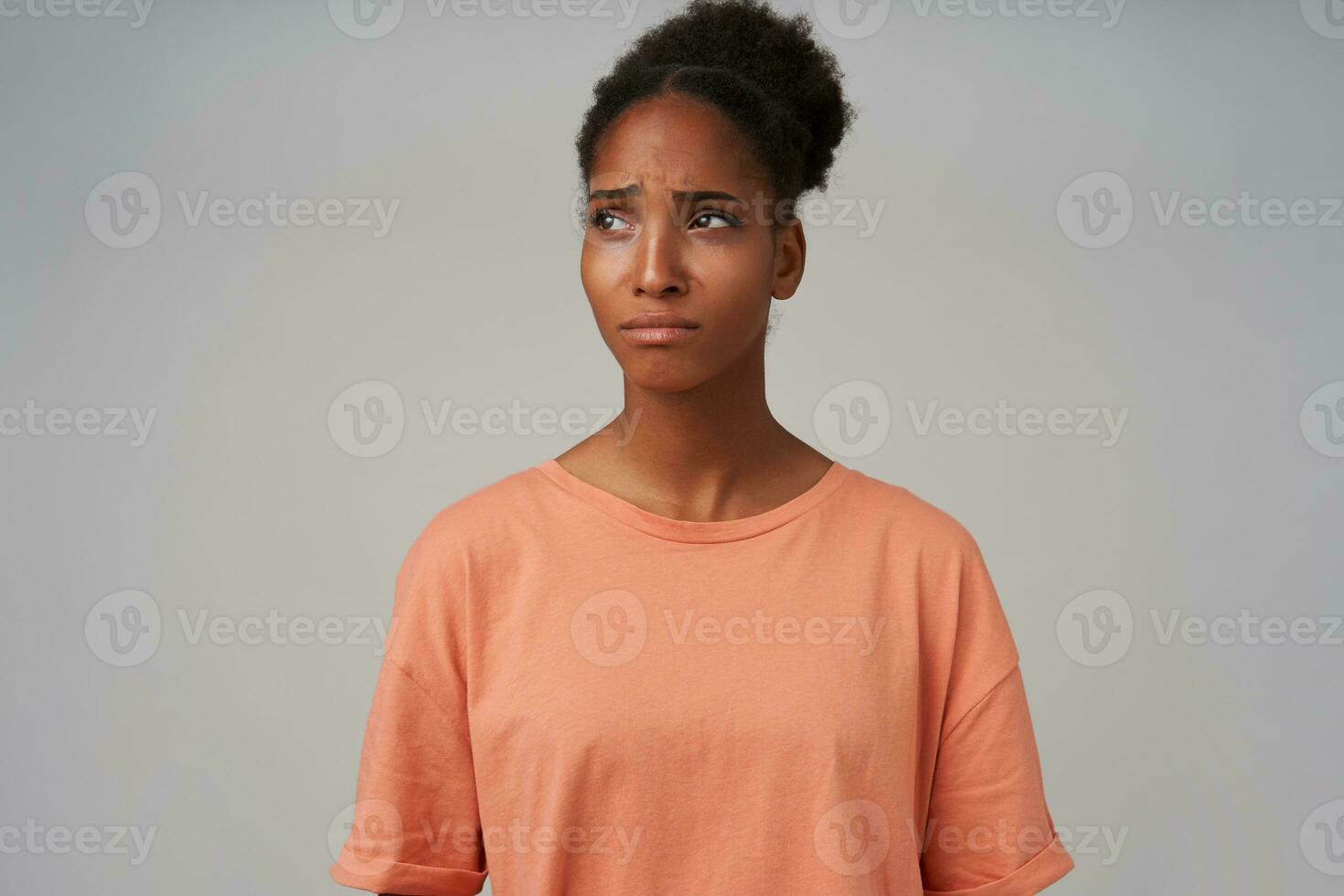 Portrait of upset young dark skinned curly lady with bun hairstyle frowning eyebrows and keeping lips folded while looking sadly aside, isolated over grey background photo