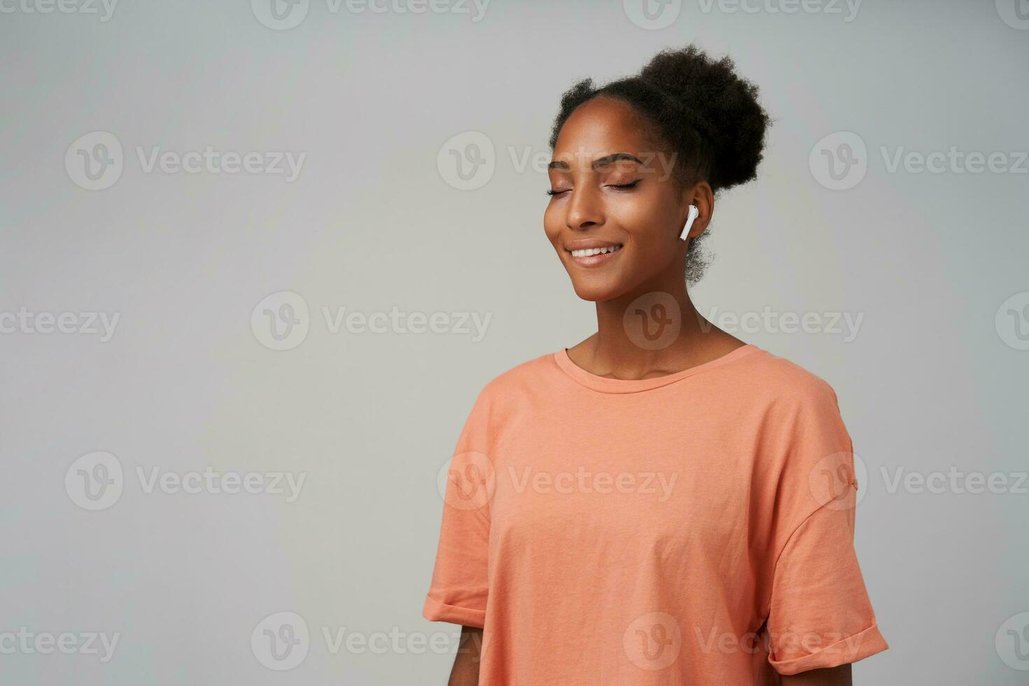Pleasant looking young lovely dark skinned curly female keeping her eyes closed while enjoying music track in her earpieces, standing over grey background photo