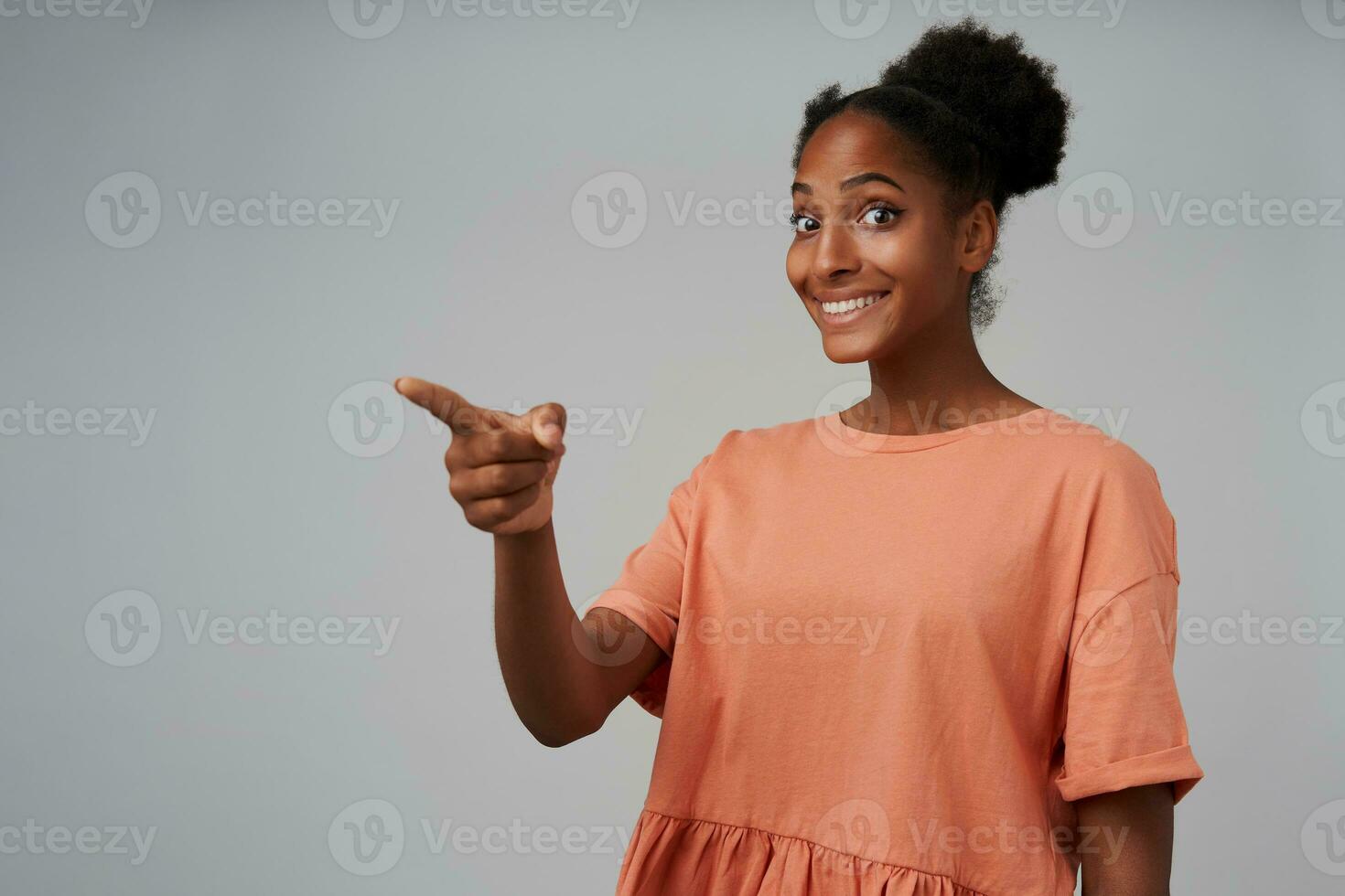 Agitated young pretty dark skinned brunette woman rounding surprisedly her eyes while showing ahead and smiling widely, isolated over grey background photo