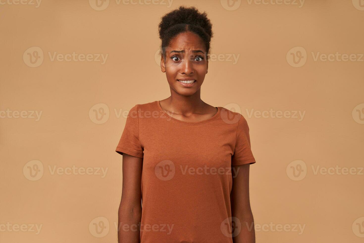 Frightened young pretty dark skinned brunette lady wearing her curly hair in knot while posing over beige background with hands down, grimacing confusedly her face while looking at camera photo