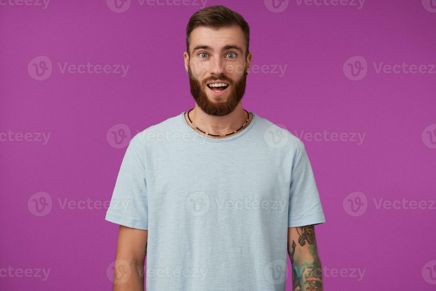 Surprised young attractive bearded man with tattooes wearing blue t-shirt while posing over purple background, keeping hands down and looking at camera with wide opened eyes photo