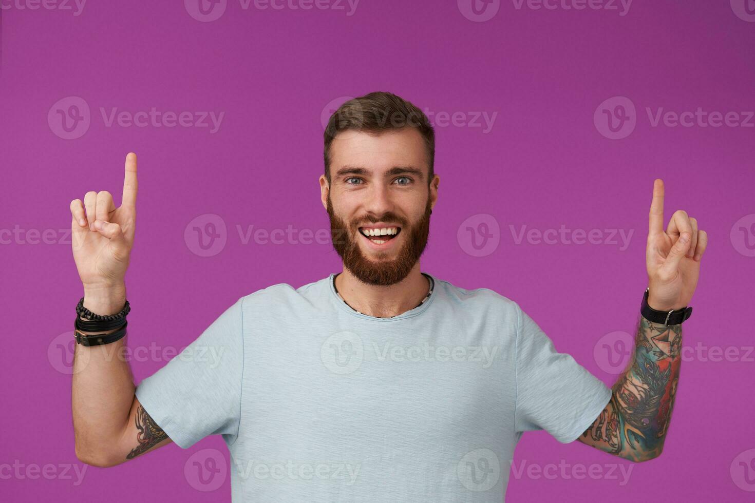 Joyful young unshaved tattooed man with short haircut wearing blue t-shirt while standing over purple background, looking at camera with wide happy smile and raising up his index fingers photo