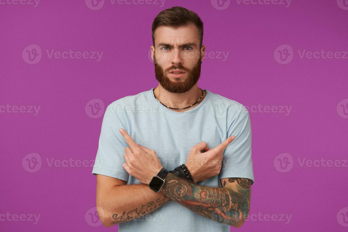 Severe bearded tattooed brunette guy with trendy haircut crossing arms on his chest and showing forefingers, looking to camera seriously and folding lips, isolated over purple background photo