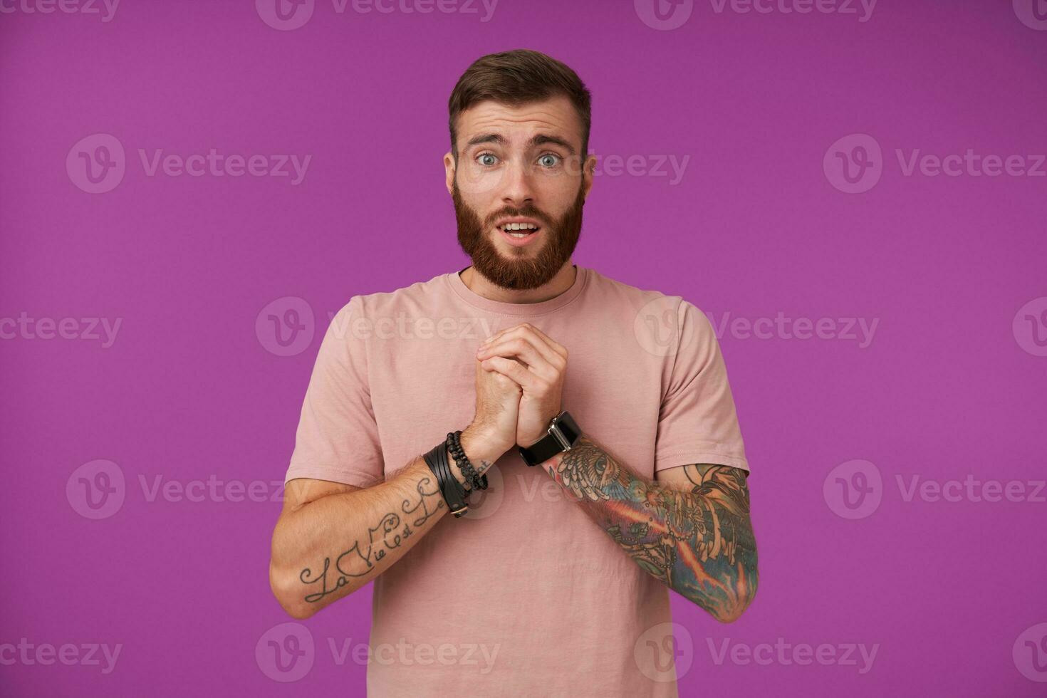 Portrait of pretty blue-eyed bearded brunette guy with tattooes raising folded hands and looking to camera pitifully, wearing beige t-shirt and trendy accessories while posing over purple background photo