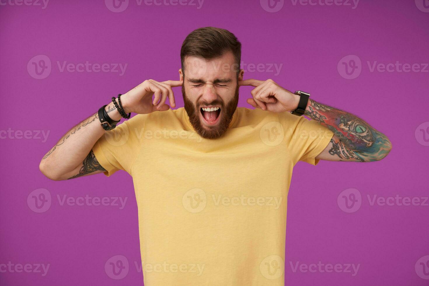 Studio shot of stressed young tattooed unshaved male covering his ears because of loud noise, keeping eyes closed and openenig mouth widely, frowning his face while standing over purple background photo