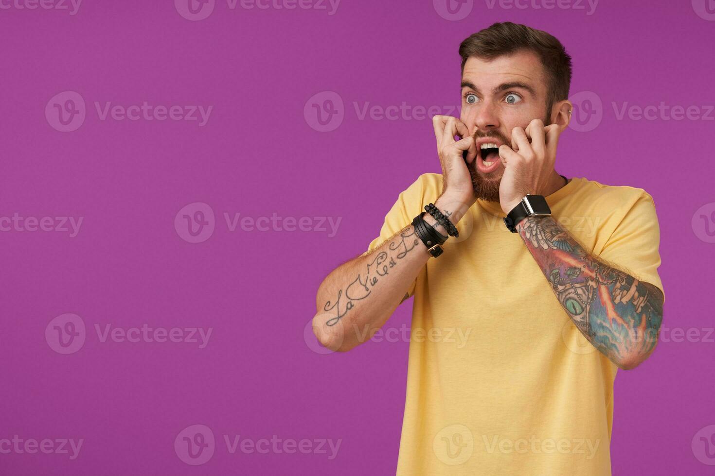 Scared young bearded man with tattooes and short haircut wearing yellow t-shirt while posing over purple background, looking aside frightened and holding his face with hands photo
