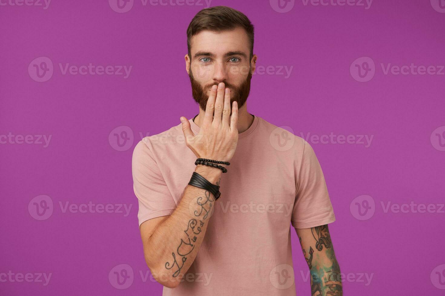 Charming young tattooed blue-eyed man with tattooes wearing beige t-shirt and trendy accessories while posing over purple background, keeping palm on his mouth and looking positively at camera photo