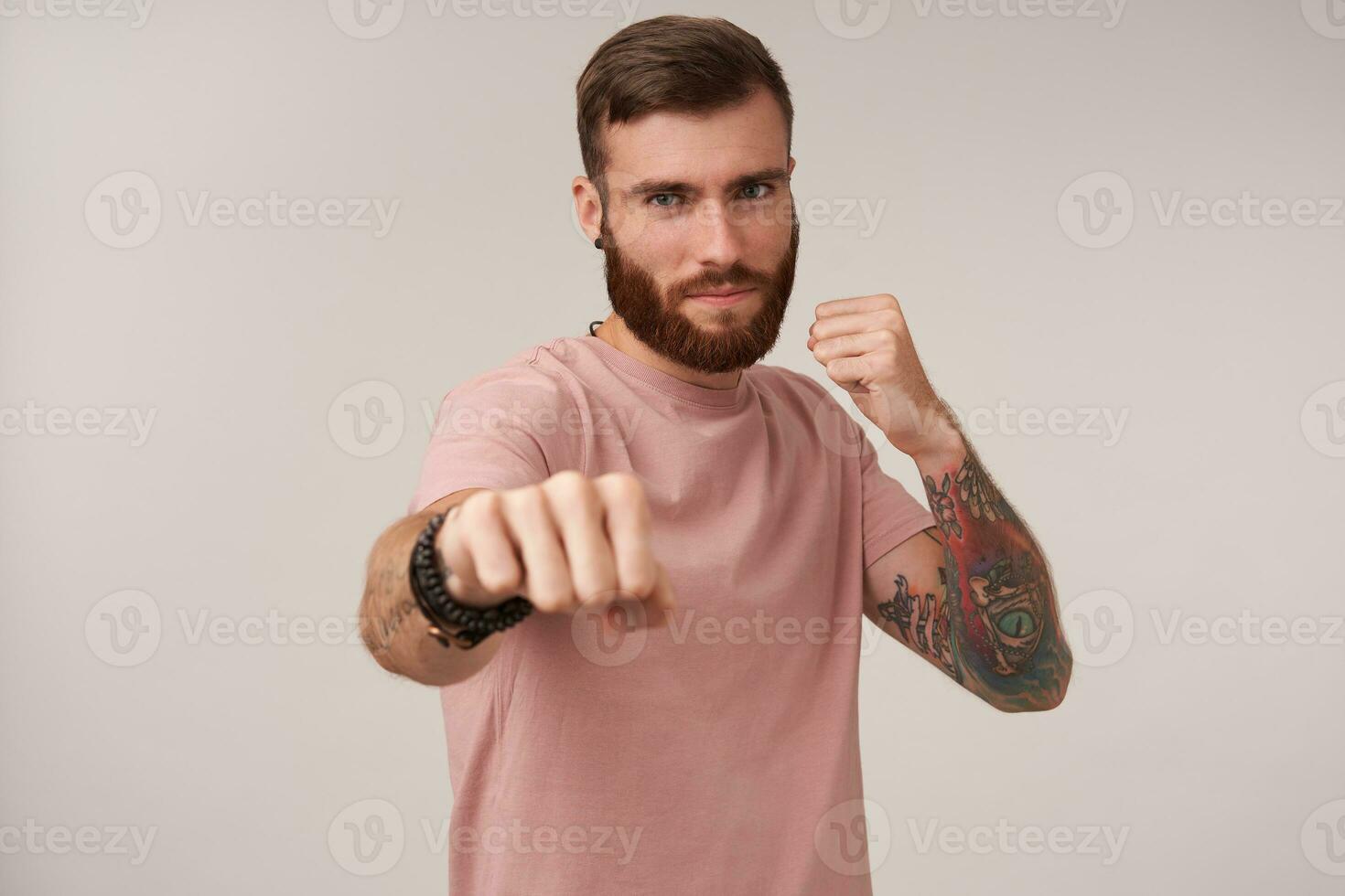 Attractive young tattooed brunette man with beard raising fist and looking to camera menacingly, being read to fight, posing over white background in casual clothes photo