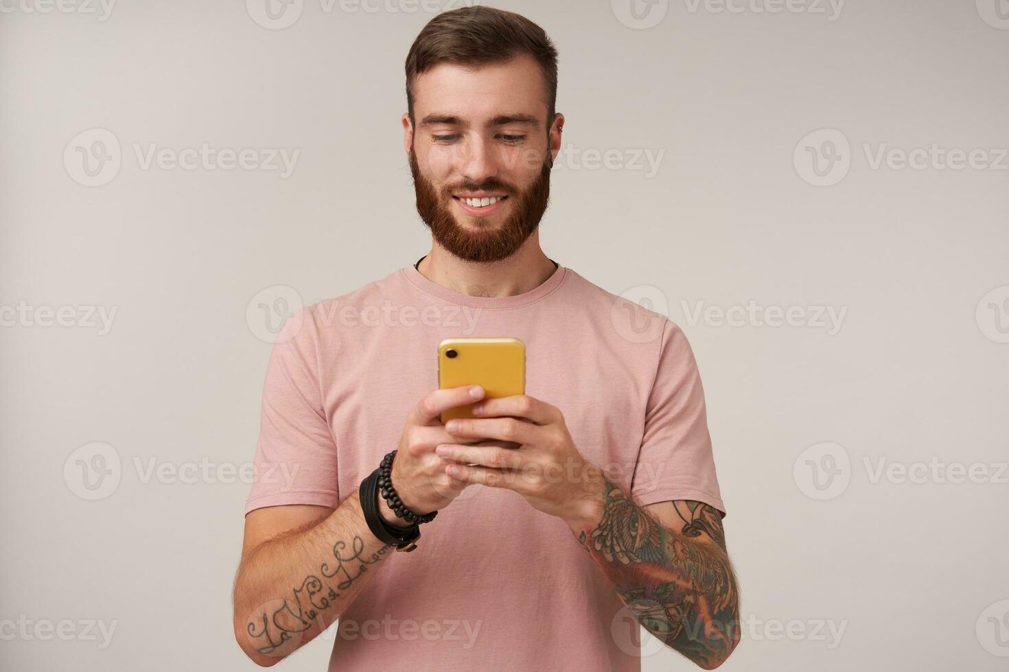 Pleasant looking young tattooed brunette male with short haircut holding smartphone in raised hands and chatting with his friends, standing over white background with sincere smile photo