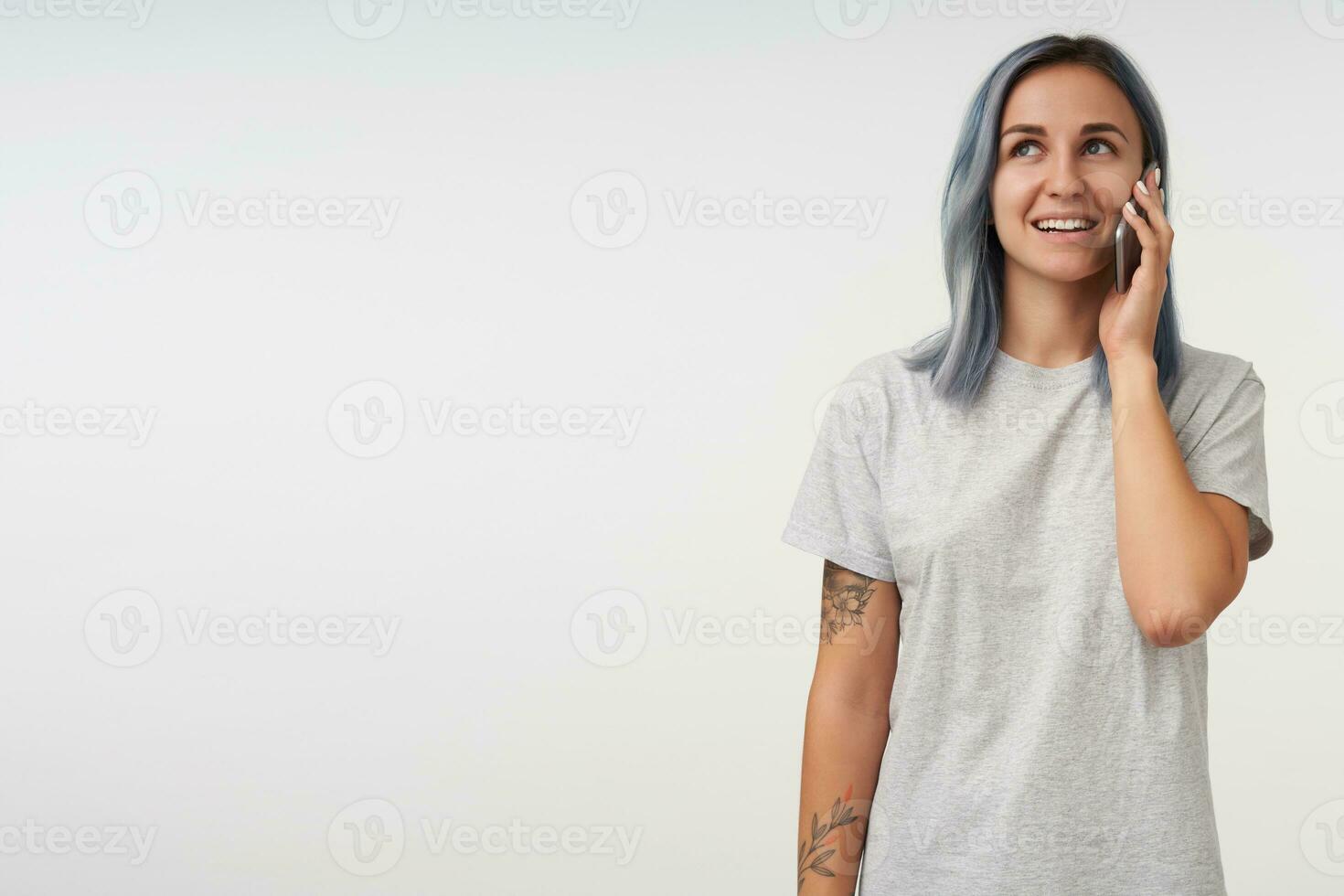 Positive young pretty tattooed lady with short blue hair smiling gently while having pleasant phone conversation, standing over white background in casual wear photo