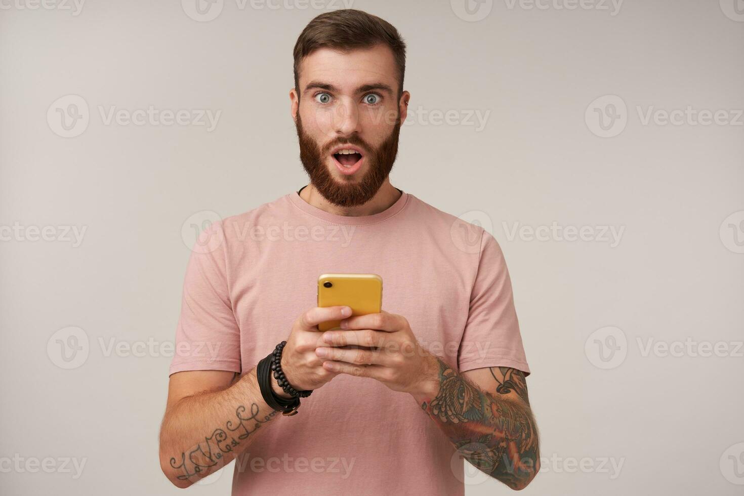 Amazed young tattooed brunette male with trendy haircut rounding his eyes and keeping mouth opened while looking at camera, wearing beige t-shirt over white background photo