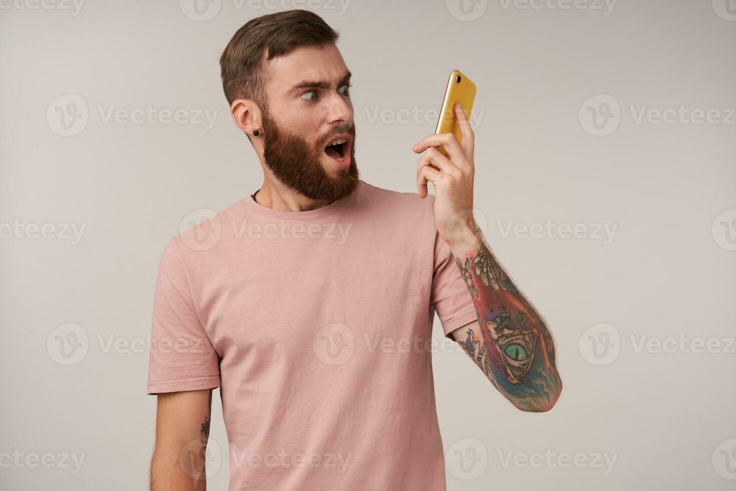 Excited young tattooed brunette man with beard looking anrgrily on handset in his hand while having tense conversation, standing over white background in beige t-shirt photo