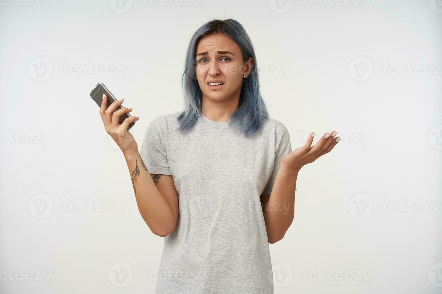 Bewildered young short haired tattooed lady with natural makeup raising her hand and frowning confusedly eyebrows, standing over white background in casual wear photo