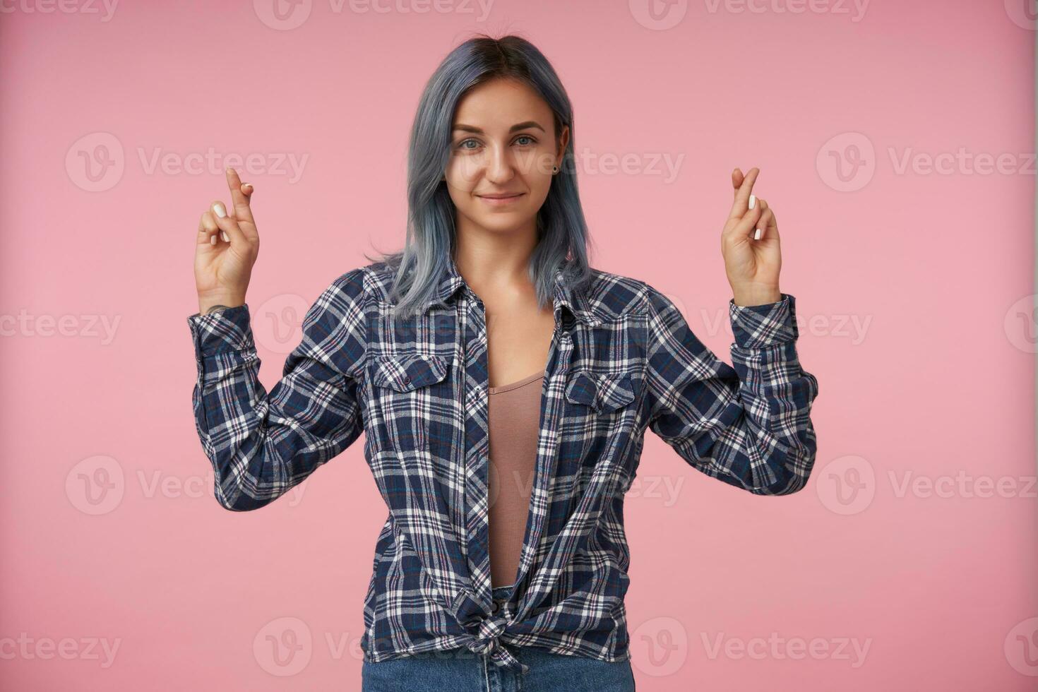 agradable mirando joven corto peludo mujer con natural maquillaje acuerdo su dedos cruzado y sonriente suavemente, vistiendo a cuadros camisa terminado rosado antecedentes foto