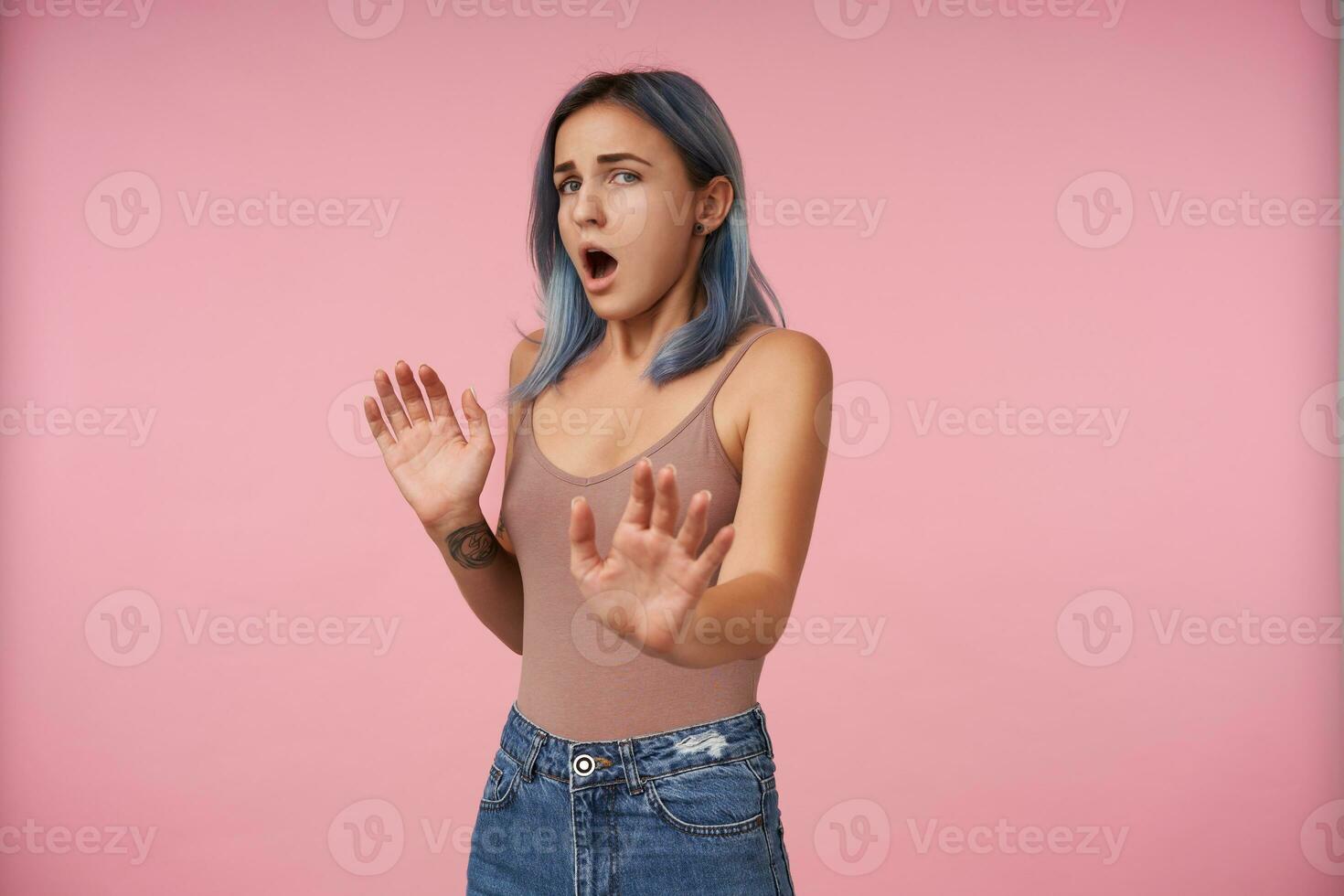 Studio photo of young pretty female with casual hairstyle raising hands in protective gesture while looking confusedly at camera, standing over pink background