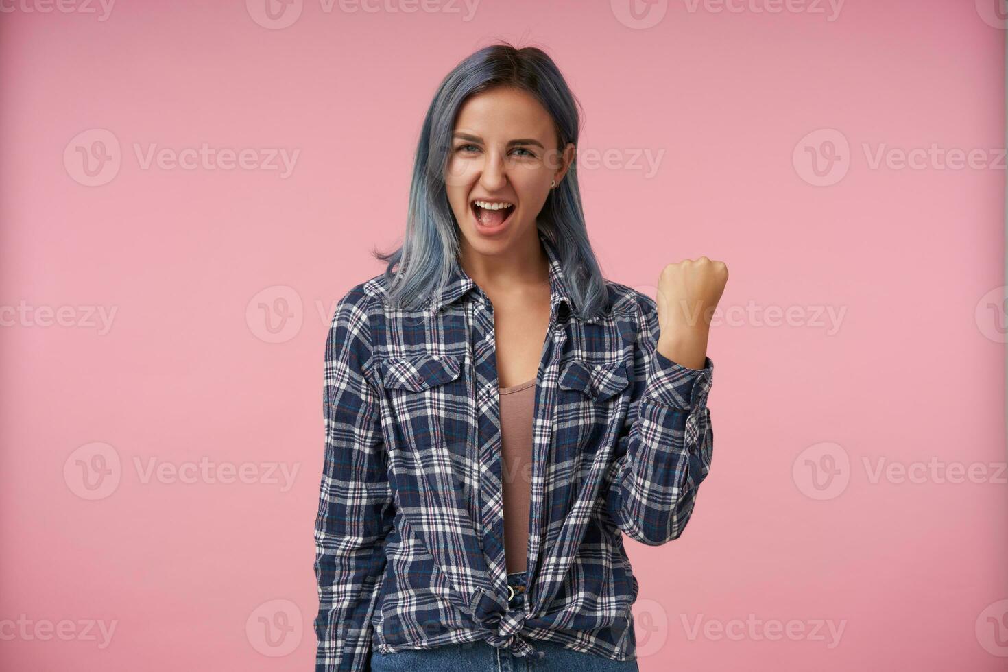 Joyful young attractive female with short blue hair raising joyfully her hand while looking exccitedly at camera, dressed in checkered shirt while posing over pink background photo