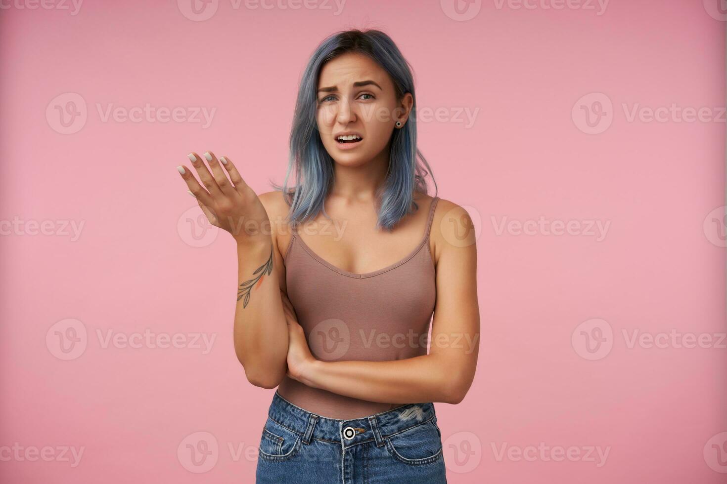Portrait of bewildered young attractive short haired woman with tattoos raising confusedly palm while looking perplexedly at camera, isolated over pink background photo