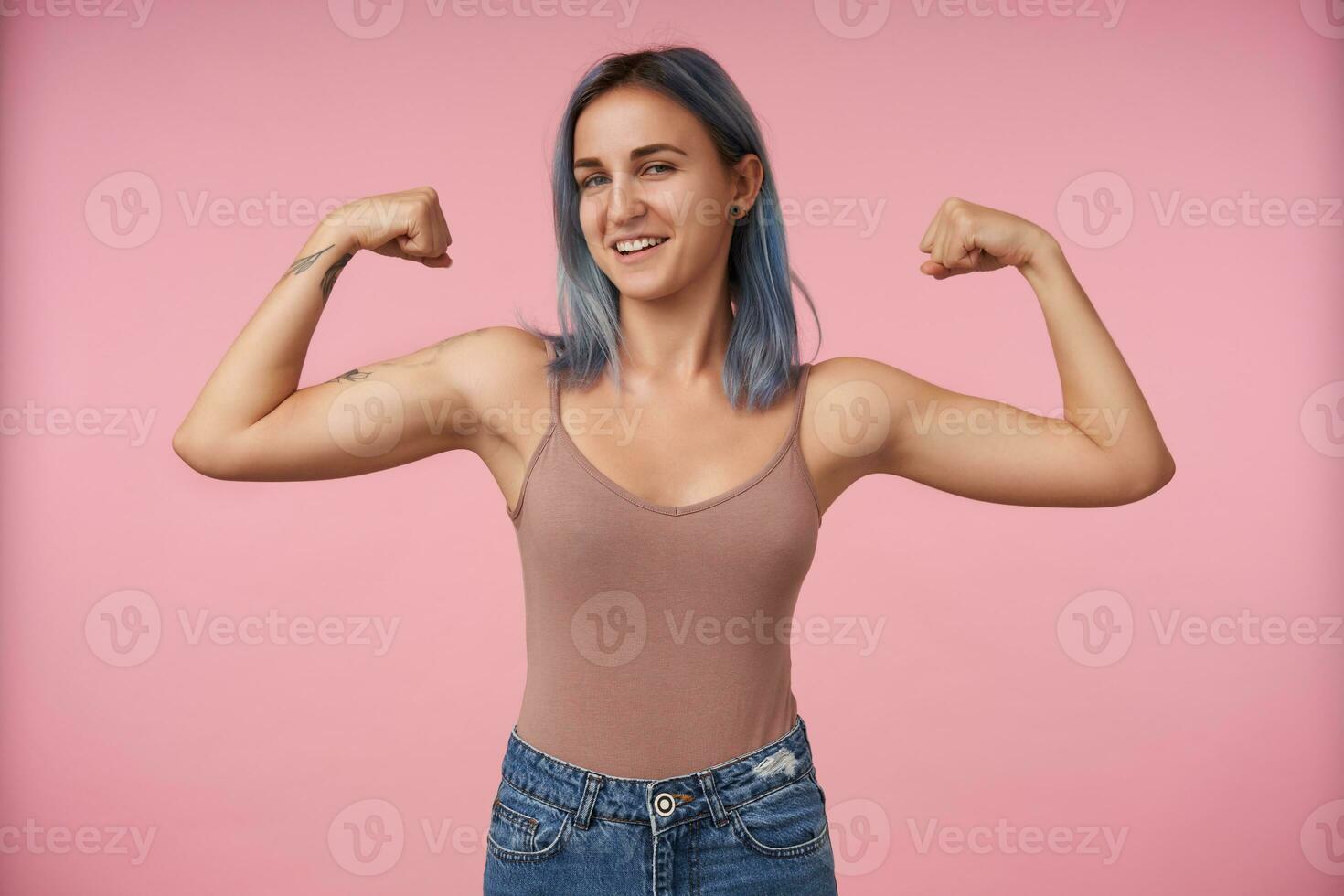 Self-confident young pretty short haired female with tattoos keeping her hands raised while demonstrating her power and looking cunningly at camera, isolated over pink background photo