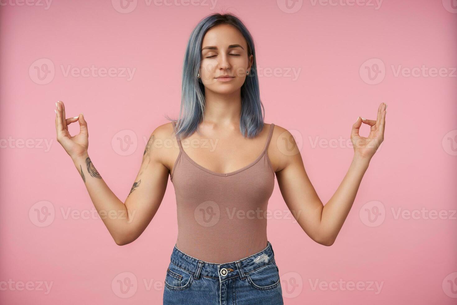 Pleasant looking young attractive blue haired female with tattoes raising hands with mudra gesture and keeping her eyes closed, standing over pink background photo